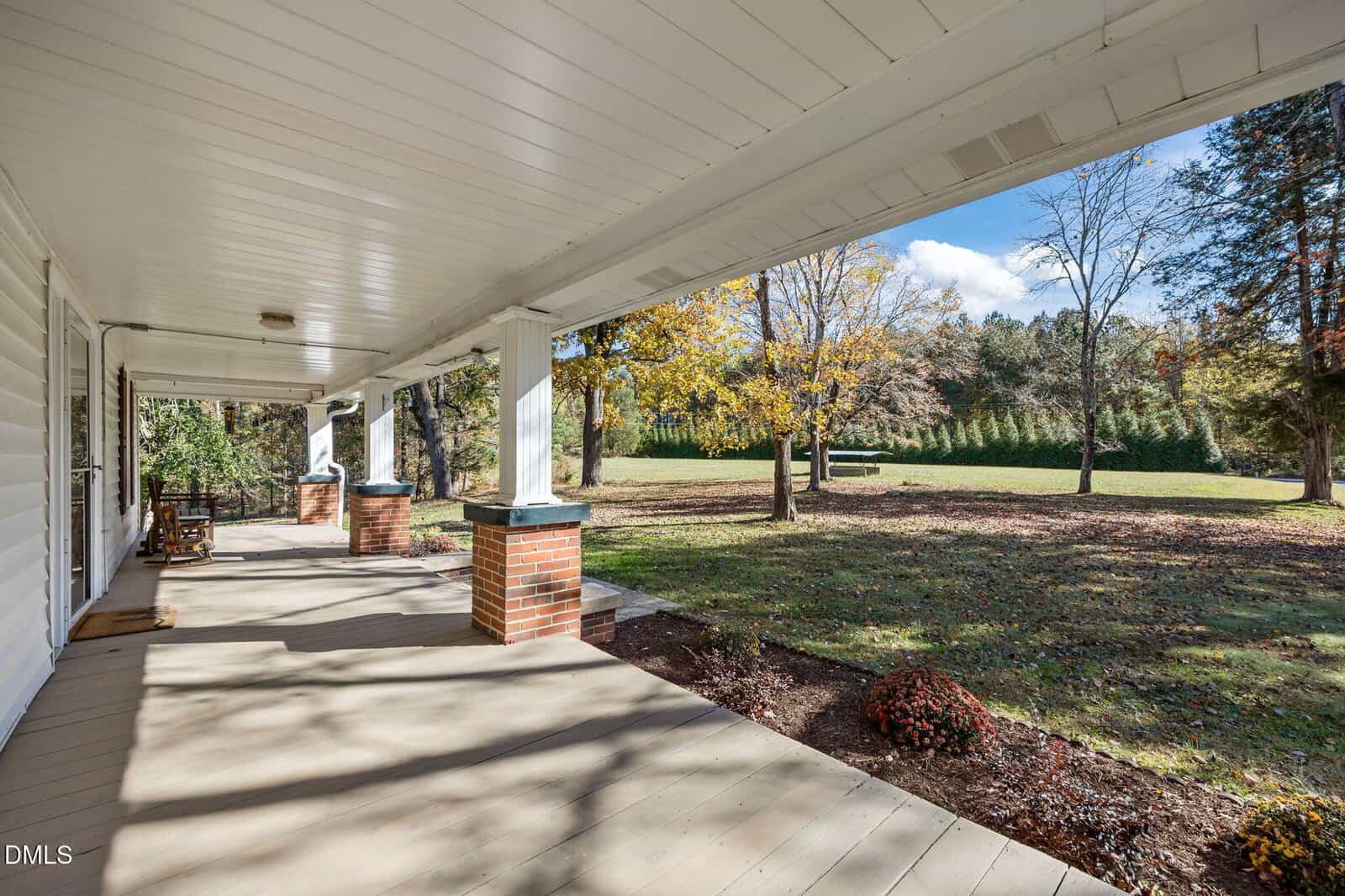 A spacious covered porch with white columns and brick bases overlooks a grassy yard with trees and a blue sky.