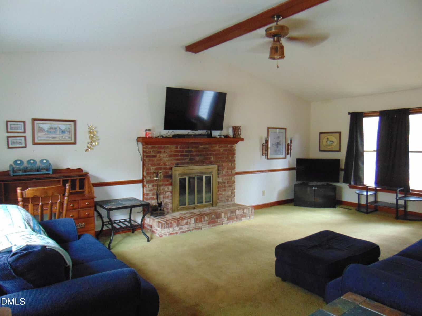 A living room with a brick fireplace, a TV mounted above it, a wooden desk to the left, a blue sofa and ottoman, two windows with dark curtains, and framed pictures on white walls.