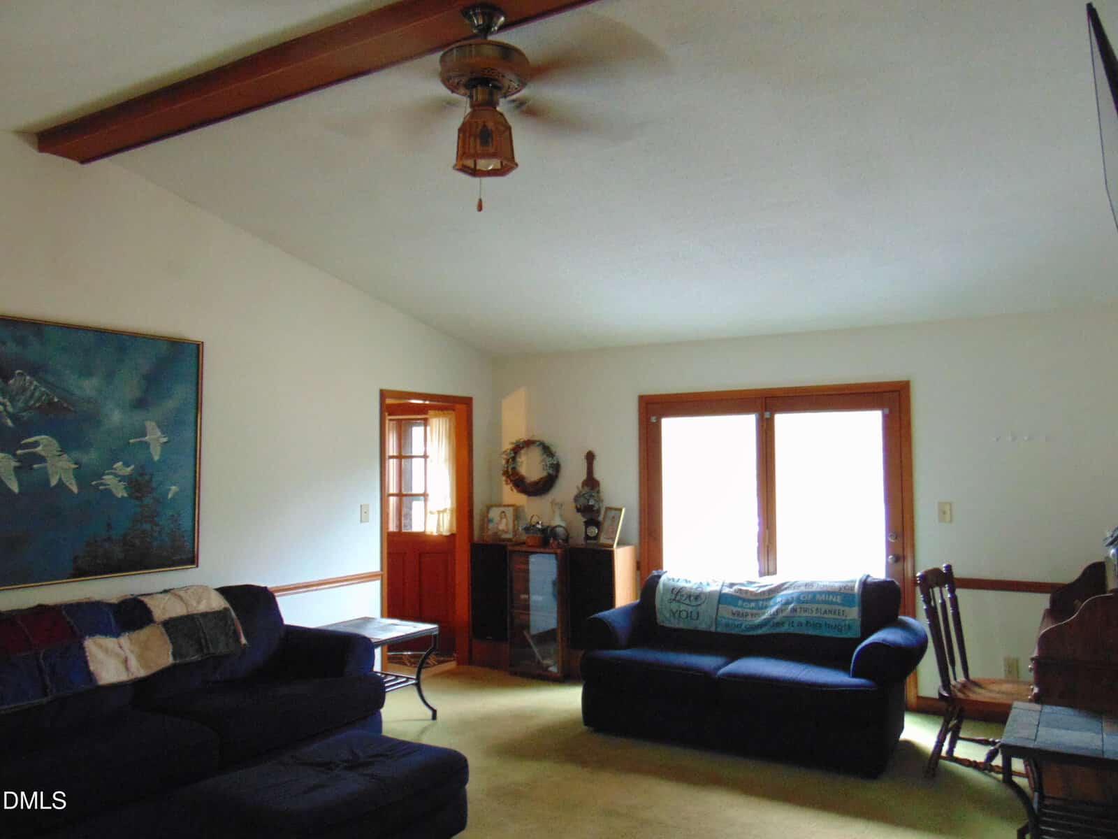 A living room with two dark blue couches, a wooden rocking chair, a ceiling fan with a light, a large painting of birds on the wall, and glass doors letting in natural light. A side table and small cabinet are also visible.