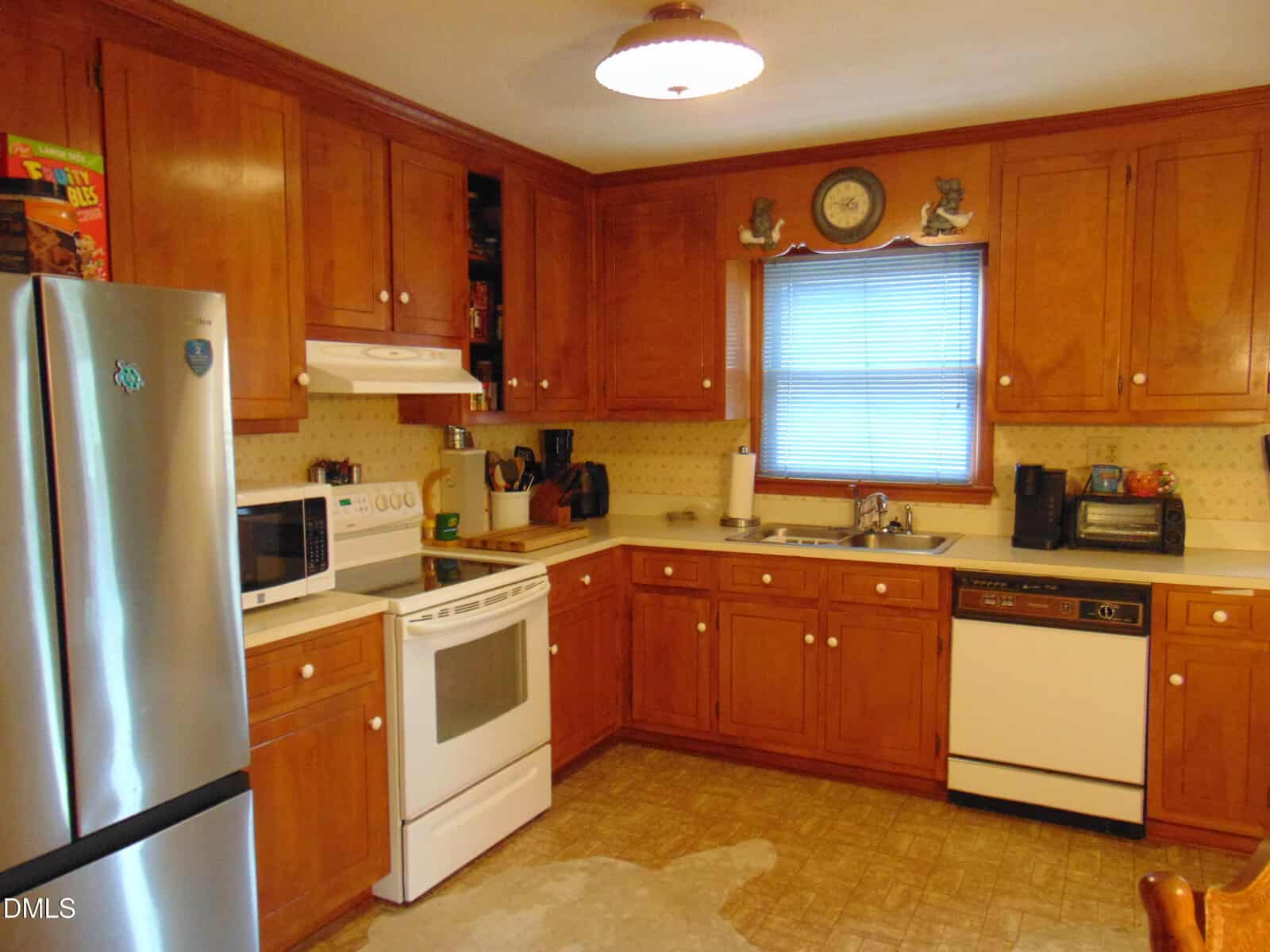 Cozy kitchen with wooden cabinets, white appliances, a stainless steel fridge, a window above the sink, various small appliances, and a light brown patterned floor.