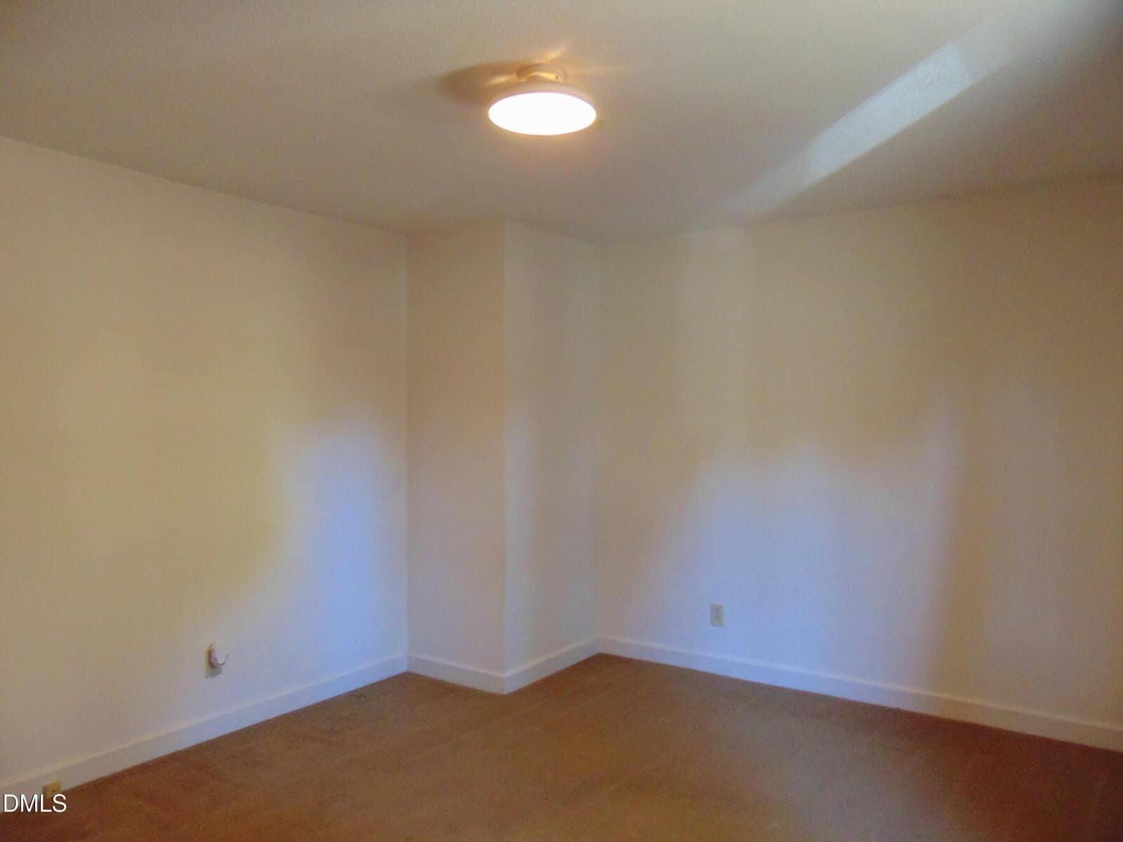 An empty room with beige walls, a ceiling light, a hardwood floor, and two electrical outlets near the baseboards. Light enters from an unseen window, creating soft shadows on the walls.