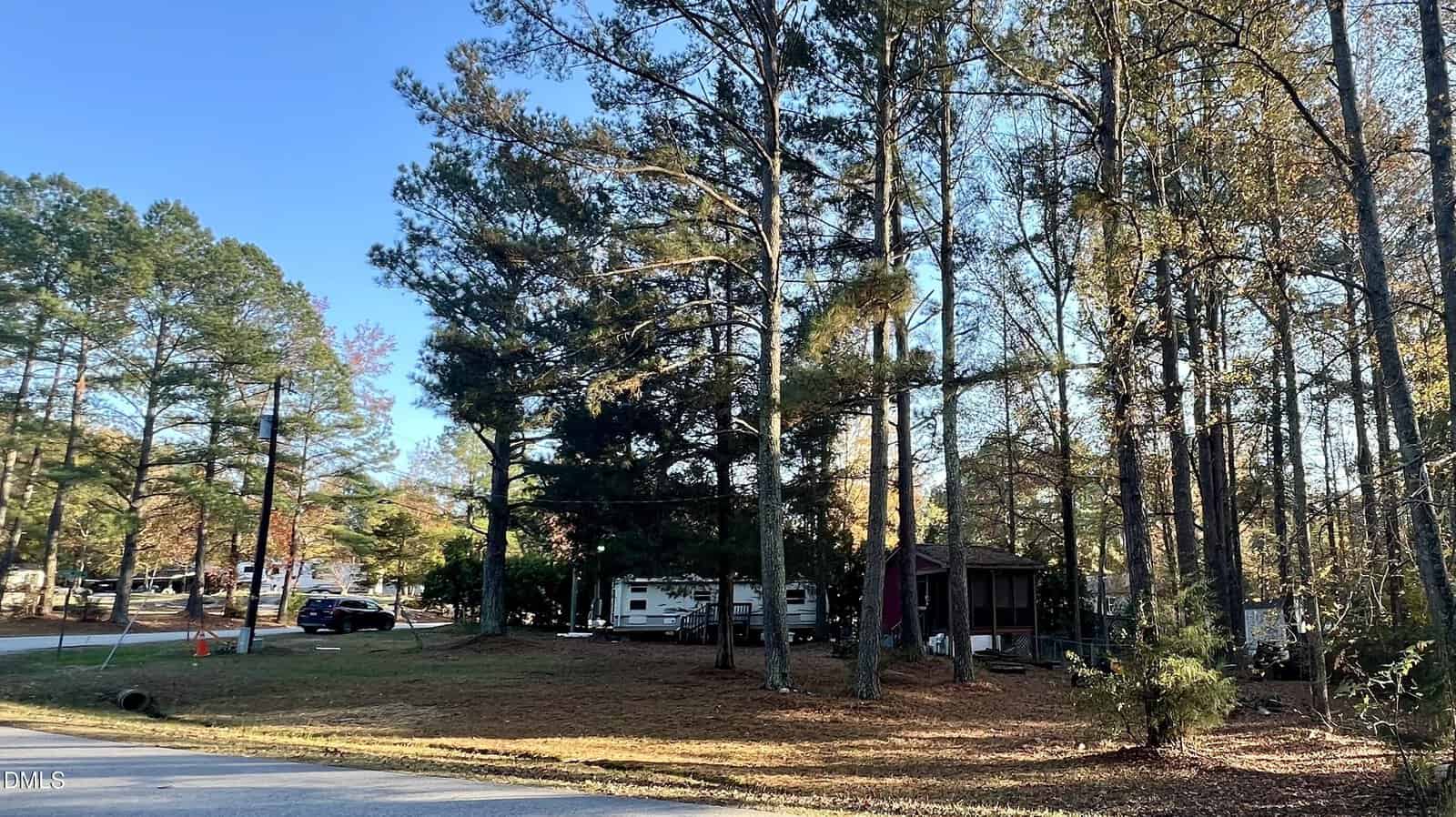 Nestled among tall trees in Louisburg, this wooded RV park at 1499 1501 Sagamore Drive features several trailers and sheds surrounded by fallen leaves, with a paved road in the foreground under a clear blue sky.