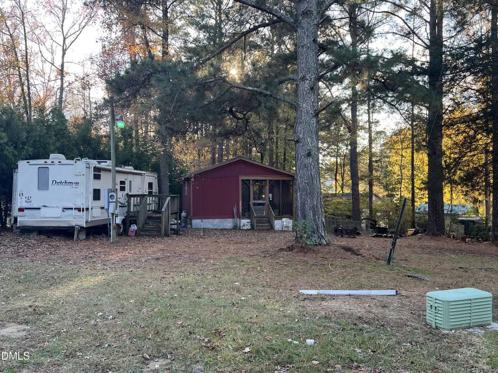 A red cabin with a screened porch sits among tall trees. A white Dutchmen RV is parked nearby, and the ground is covered with brown pine needles. Utility boxes and yard items are scattered in the grassy foreground.