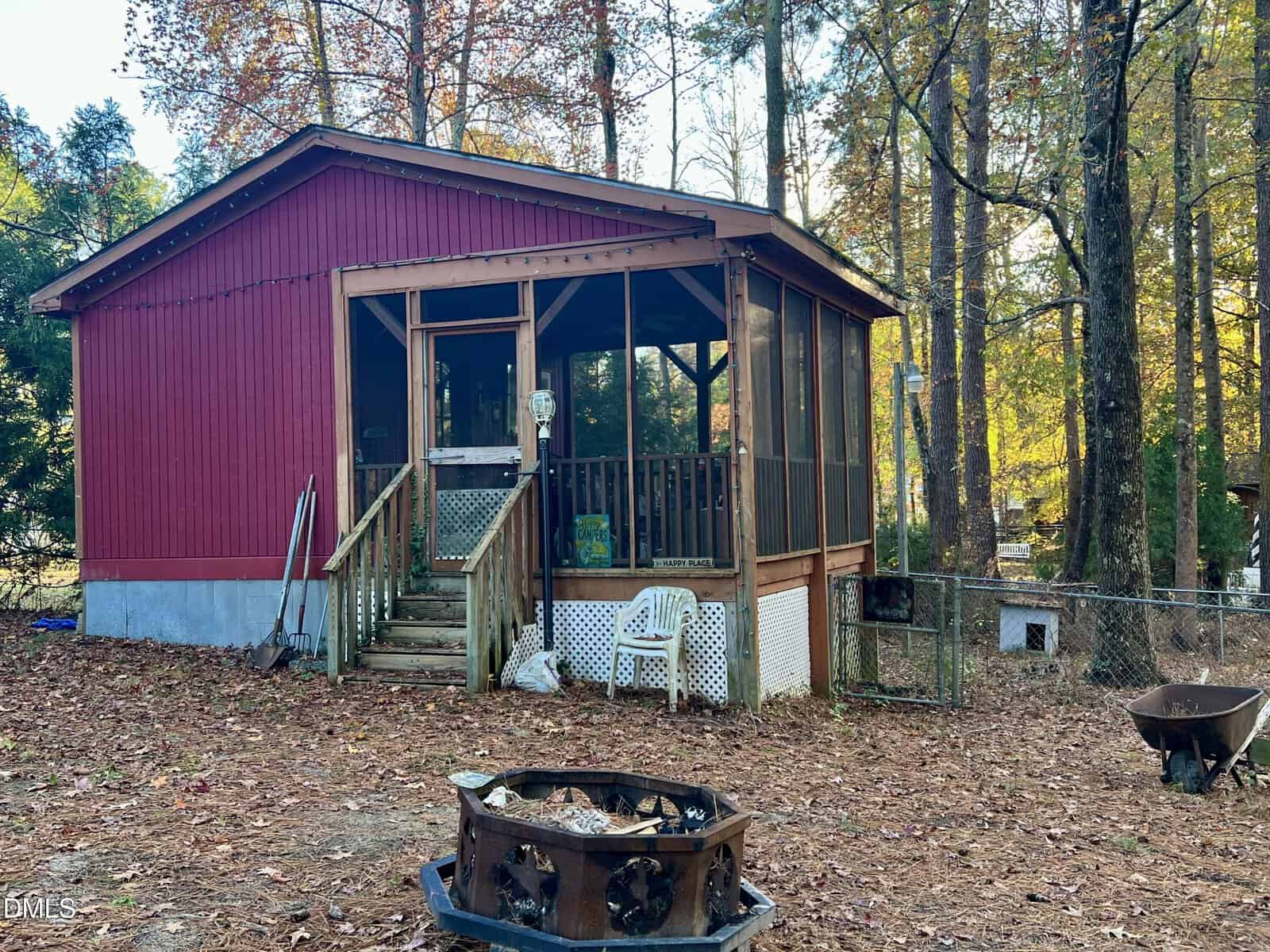 A small red cabin with a screened porch sits among tall trees. Leaves cover the ground, outdoor chairs and tools are on the porch, and a fire pit is in the foreground. A chain-link fence borders the yard.