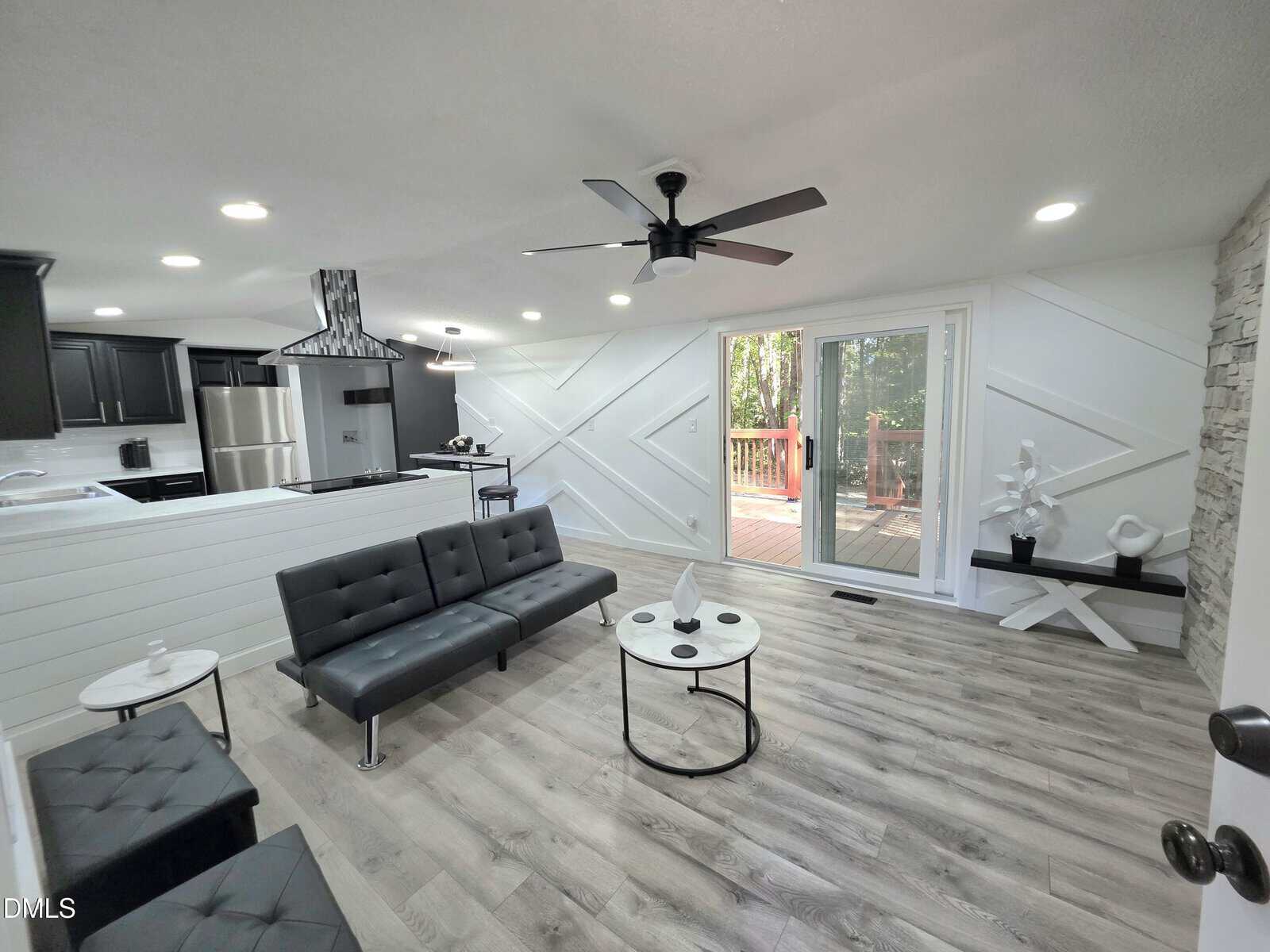 Modern living room with black leather sofa, round coffee tables, light wood flooring, and a ceiling fan. Sliding glass doors open to a deck, with decorative accents and a white geometric wall design. Kitchen visible in the background.
