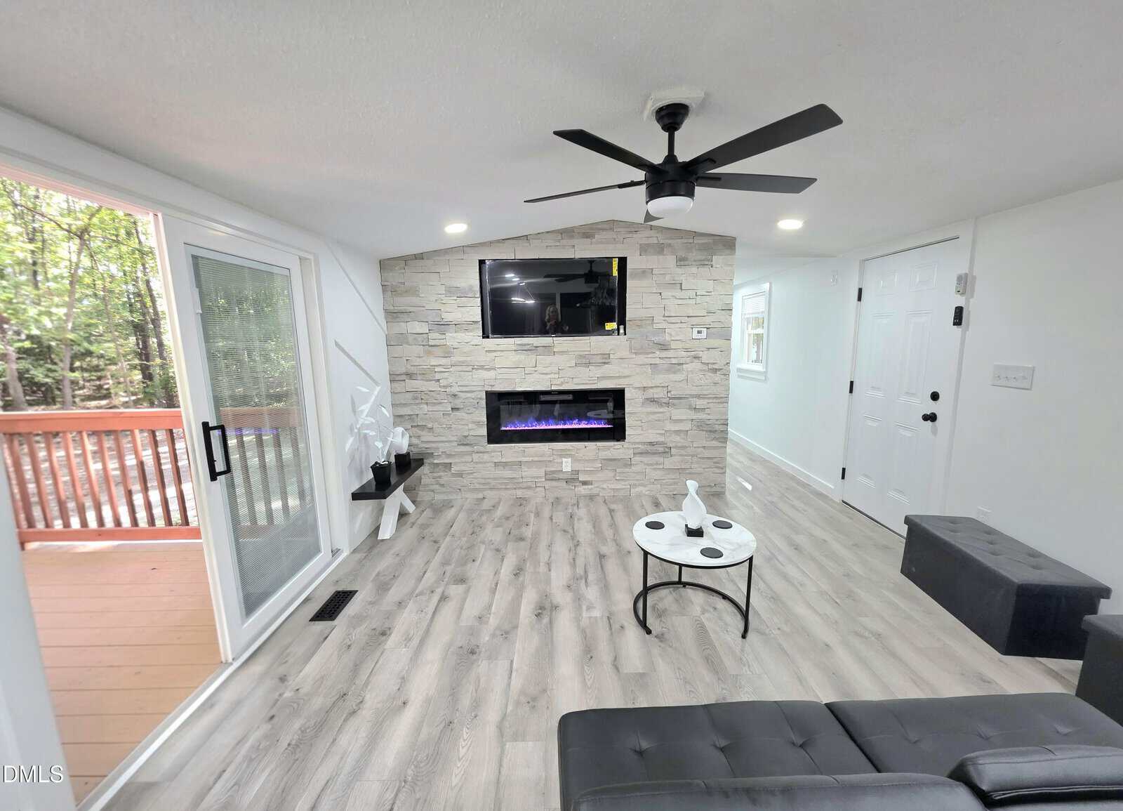 Modern living room with light wood floors, stone accent wall with mounted TV and electric fireplace, black ceiling fan, small round coffee table, sliding glass door to a deck, and a white door to the right.