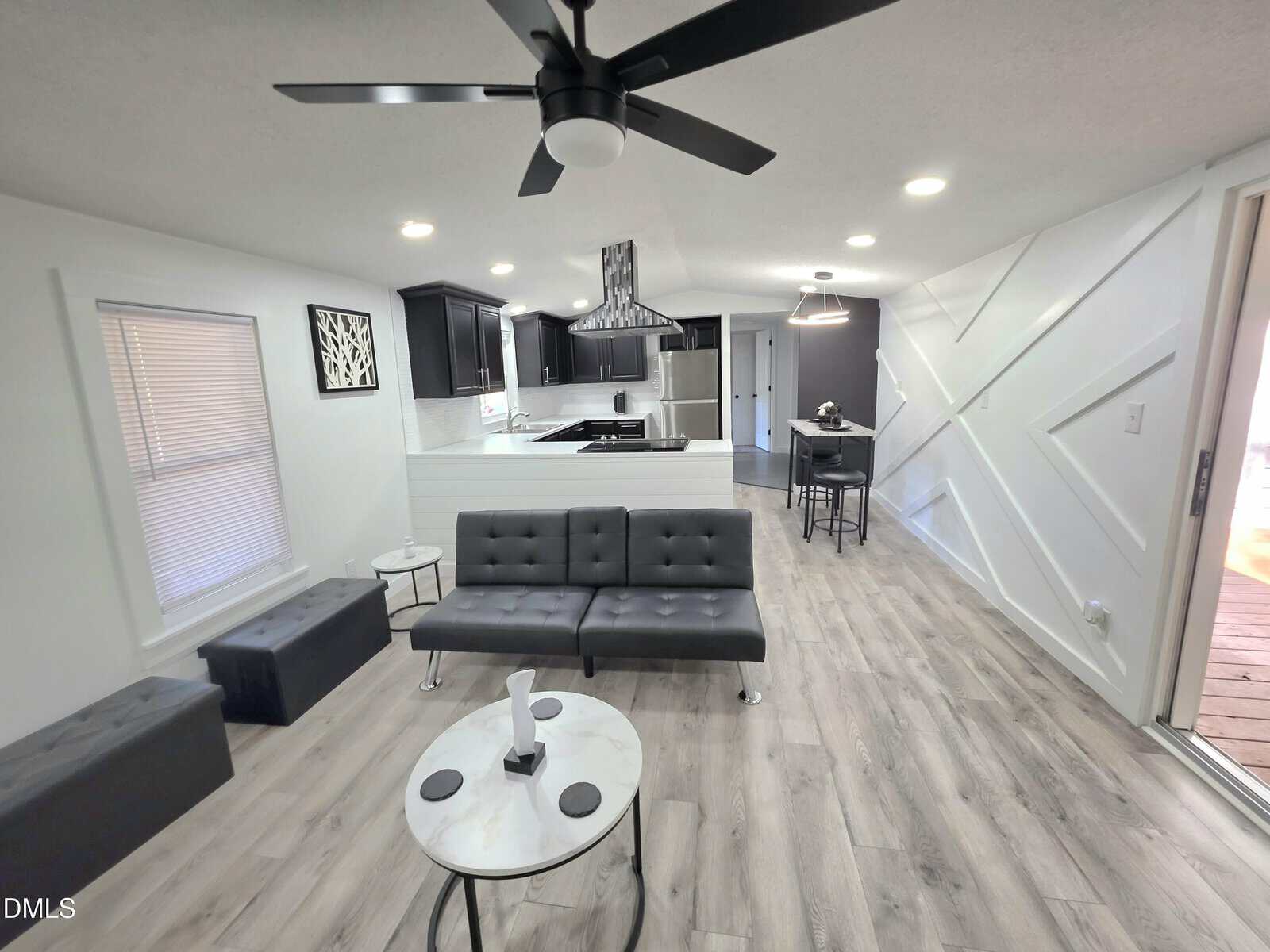 Modern living room and kitchen with gray flooring, black and white furniture, a ceiling fan, and geometric wall design. The open layout includes a sofa, coffee table, bar seating, and stainless steel appliances.
