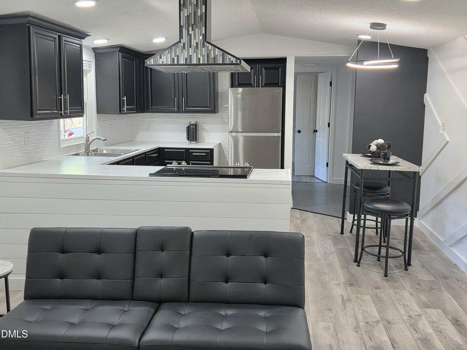 Modern open-concept kitchen and living area with black cabinets, stainless steel appliances, gray sofa, small dining table with two chairs, light wood floors, and white shiplap walls.