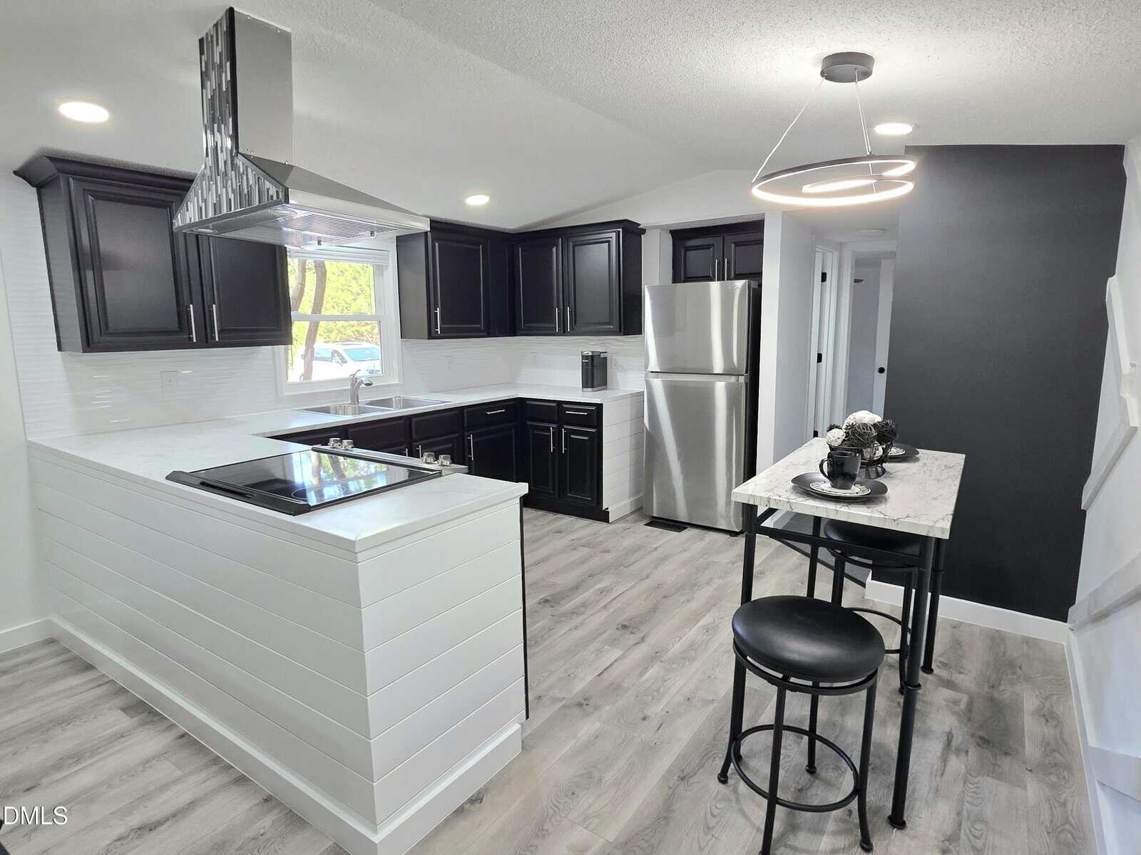 Modern kitchen with black cabinets, stainless steel appliances, light wood flooring, white walls, a small dining table with two chairs, and a large window above the sink letting in natural light.