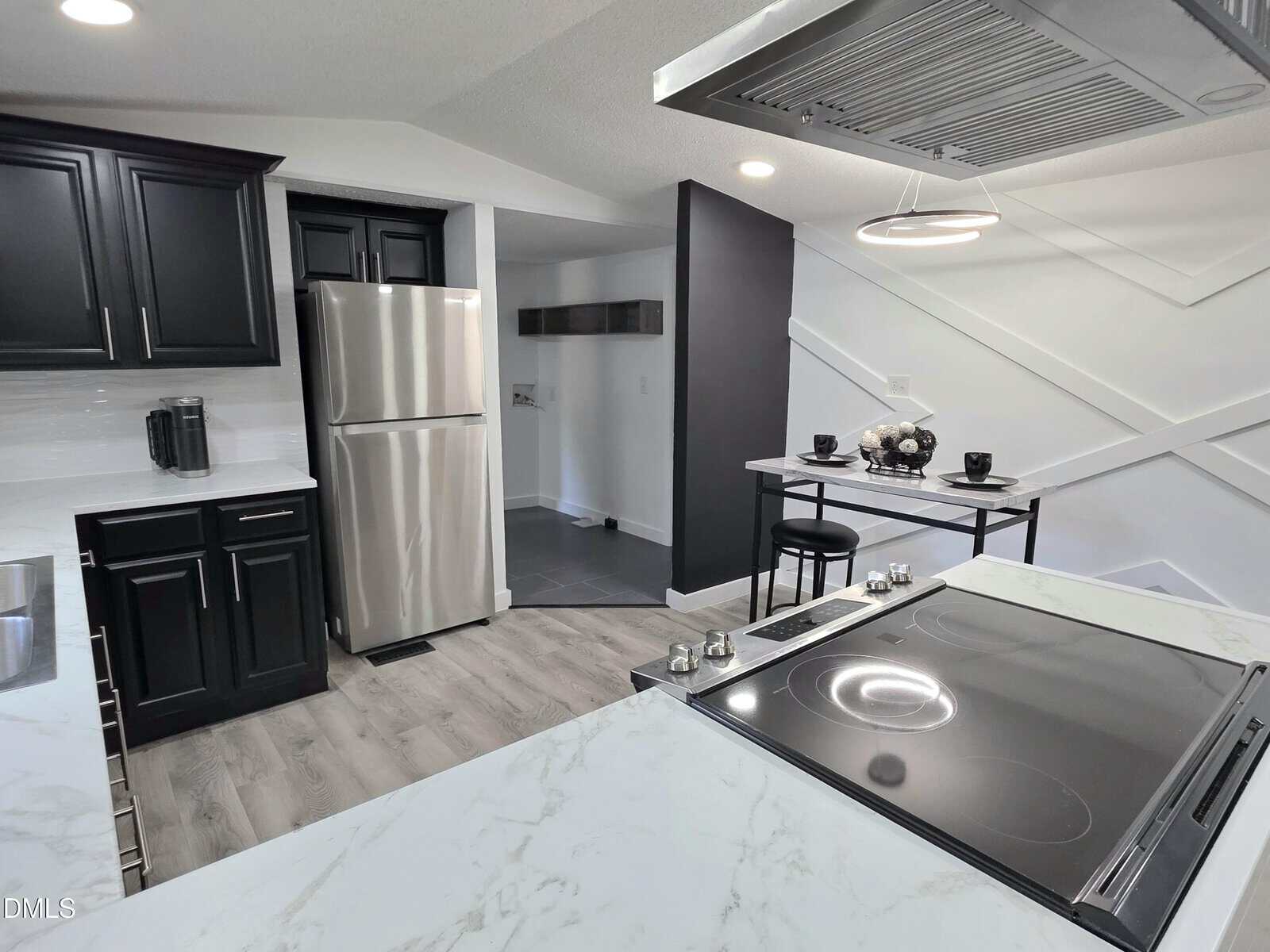 Modern kitchen with black cabinets, stainless steel appliances, a marble countertop, and a small dining area with two chairs and place settings. The space features light wood flooring and white walls with geometric accents.