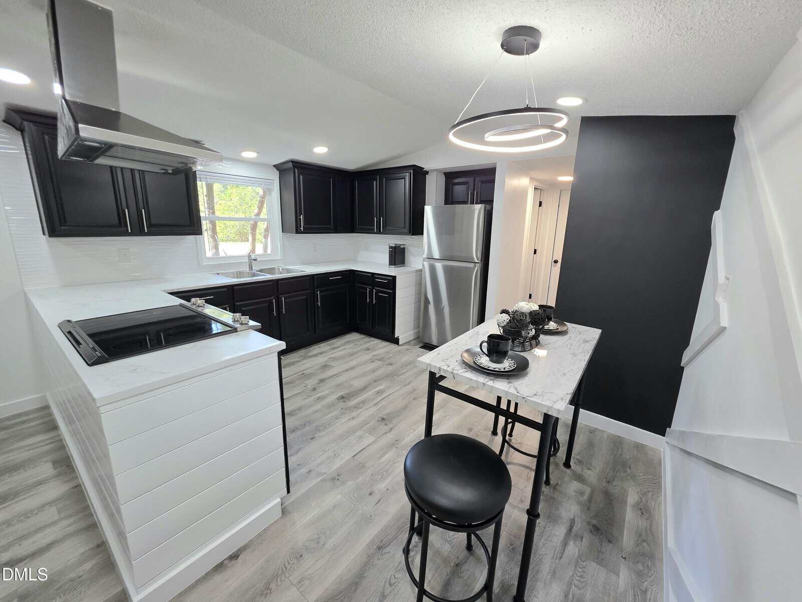 Modern kitchen with black cabinets, stainless steel appliances, light wood flooring, and a small dining table set for two. A geometric light fixture hangs above the table, and natural light enters through a window above the sink.