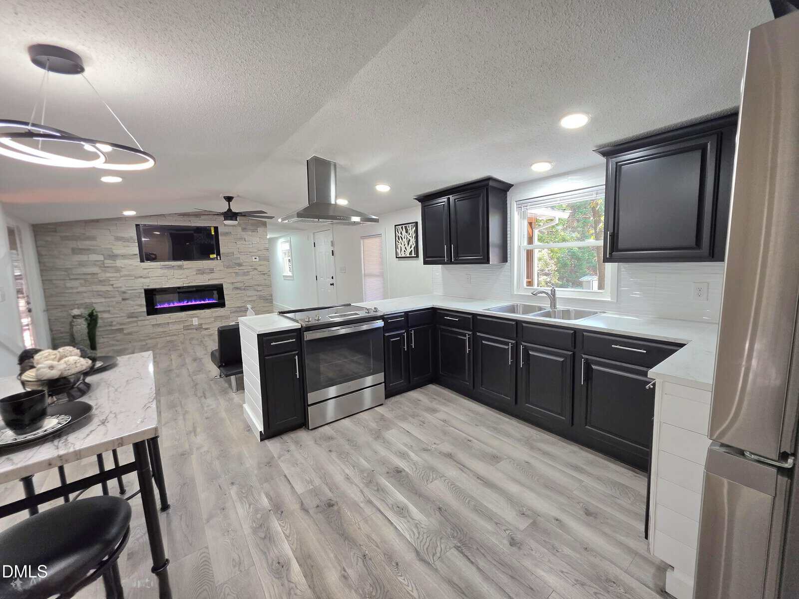 Modern kitchen with black cabinets, stainless steel appliances, white countertops, and gray wood flooring. Open layout leads to a living area with a wall-mounted TV and an electric fireplace set in a stone accent wall.