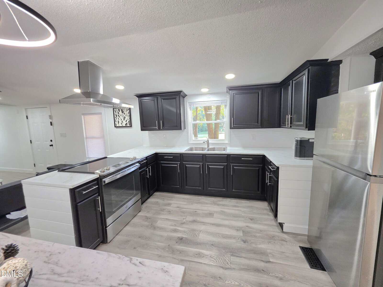 Modern kitchen with black cabinets, stainless steel appliances, and white countertops. Wooden floors and a large window above the sink let in natural light. Recessed lighting brightens the space.