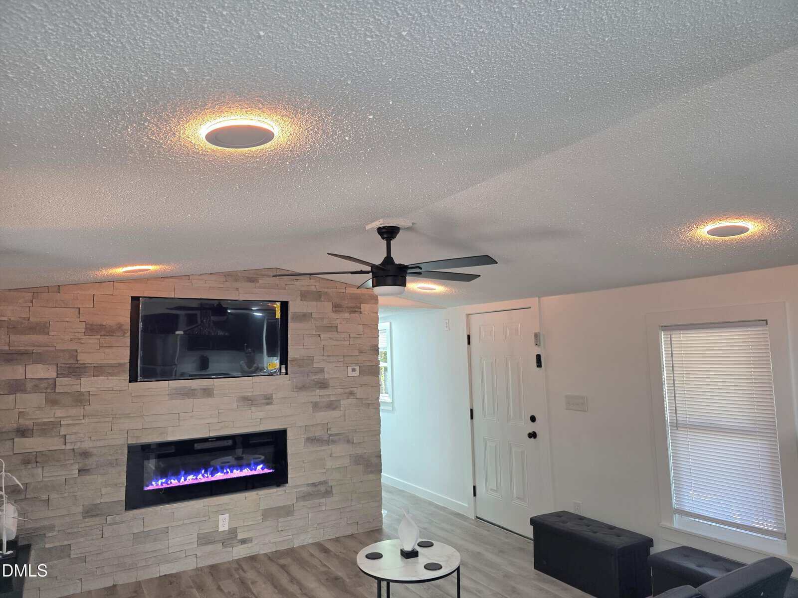 Modern living room with a stone accent wall, mounted TV, electric fireplace, ceiling fan, recessed lighting, white door, window blinds, wood floor, and small round table.