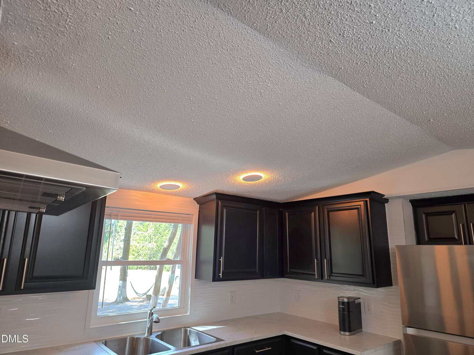 A modern kitchen with black cabinets, white countertops, stainless steel appliances, a window above the sink, and two round recessed lights in a textured ceiling.