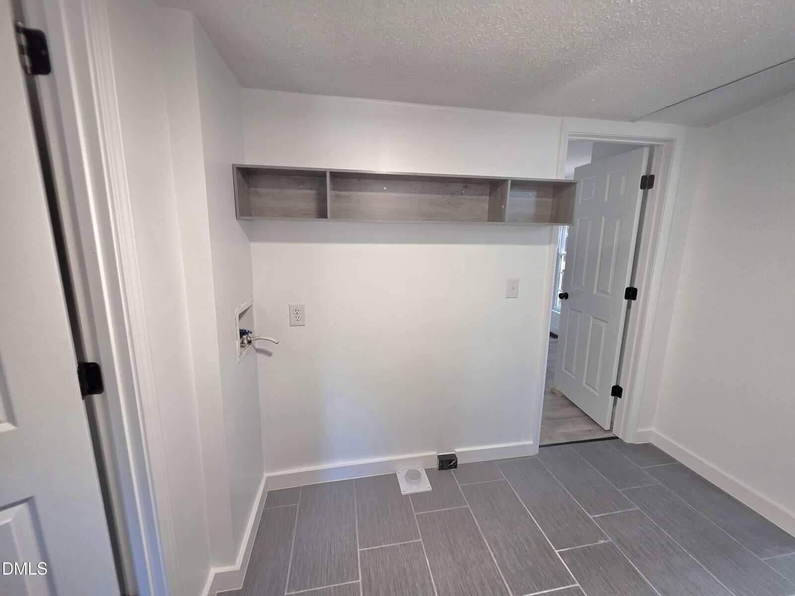 Small laundry room with gray floor tiles, a wall-mounted shelf, electrical outlets, and a water hookup. There is a white door open to an adjacent room. Walls are painted white.