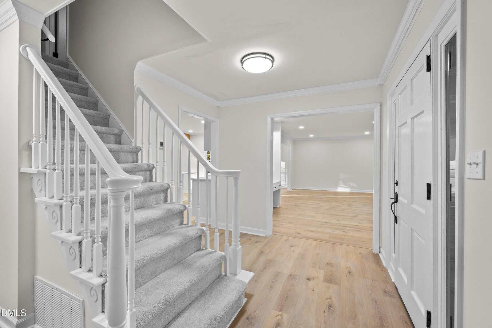 A carpeted staircase with white railings leads upward from a bright entryway featuring light wood floors, white walls, and white doors, with an adjacent open room visible.