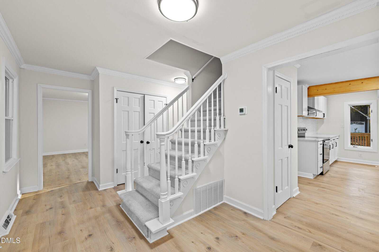 Interior view of a modern home showing a carpeted staircase with white railing, light wood flooring, white walls, and doors leading to other rooms including a visible kitchen with stainless steel appliances.