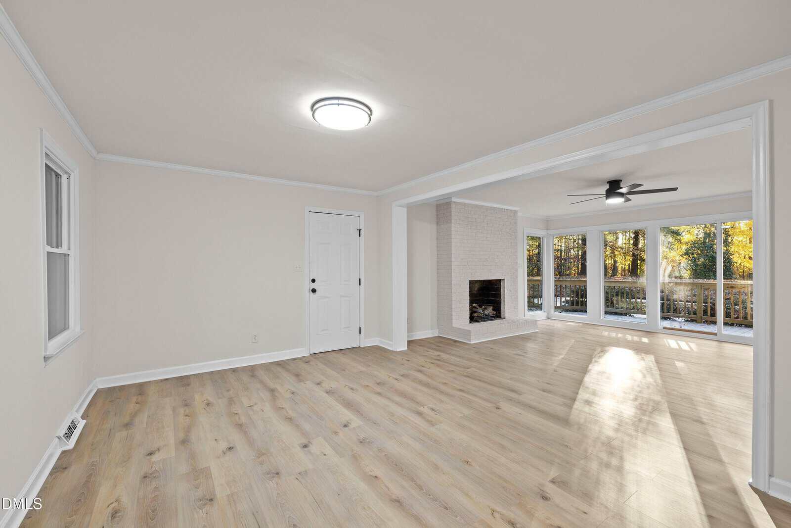 A spacious living room with light wood floors, white walls, a white front door, a fireplace, large windows, and glass doors leading to a deck with trees visible outside. The room has ceiling lights and crown molding.