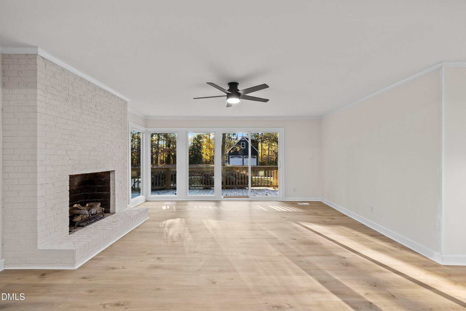 Spacious living room with light wood flooring, white brick fireplace, ceiling fan, and large windows overlooking a wooded backyard and deck. Sunlight streams through the windows, creating a bright, airy atmosphere.