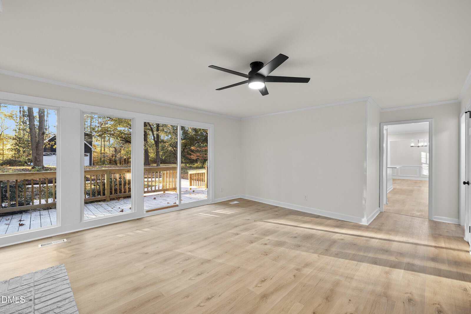 Bright living room with light wood floors, large windows and sliding glass doors leading to a deck, white walls, and a black ceiling fan. Adjacent room visible through a wide doorway.