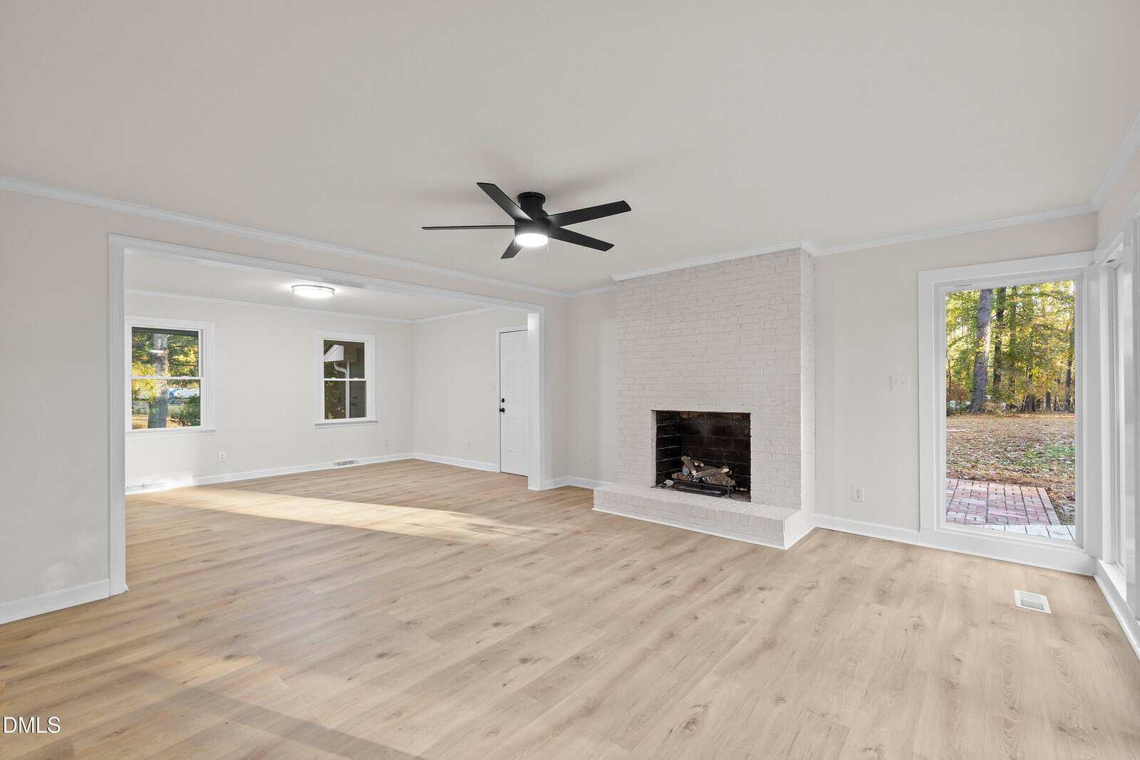 A spacious living room with light wood flooring, a white brick fireplace, white walls, and a black ceiling fan. Large windows and a glass door provide views of a wooded outdoor area.