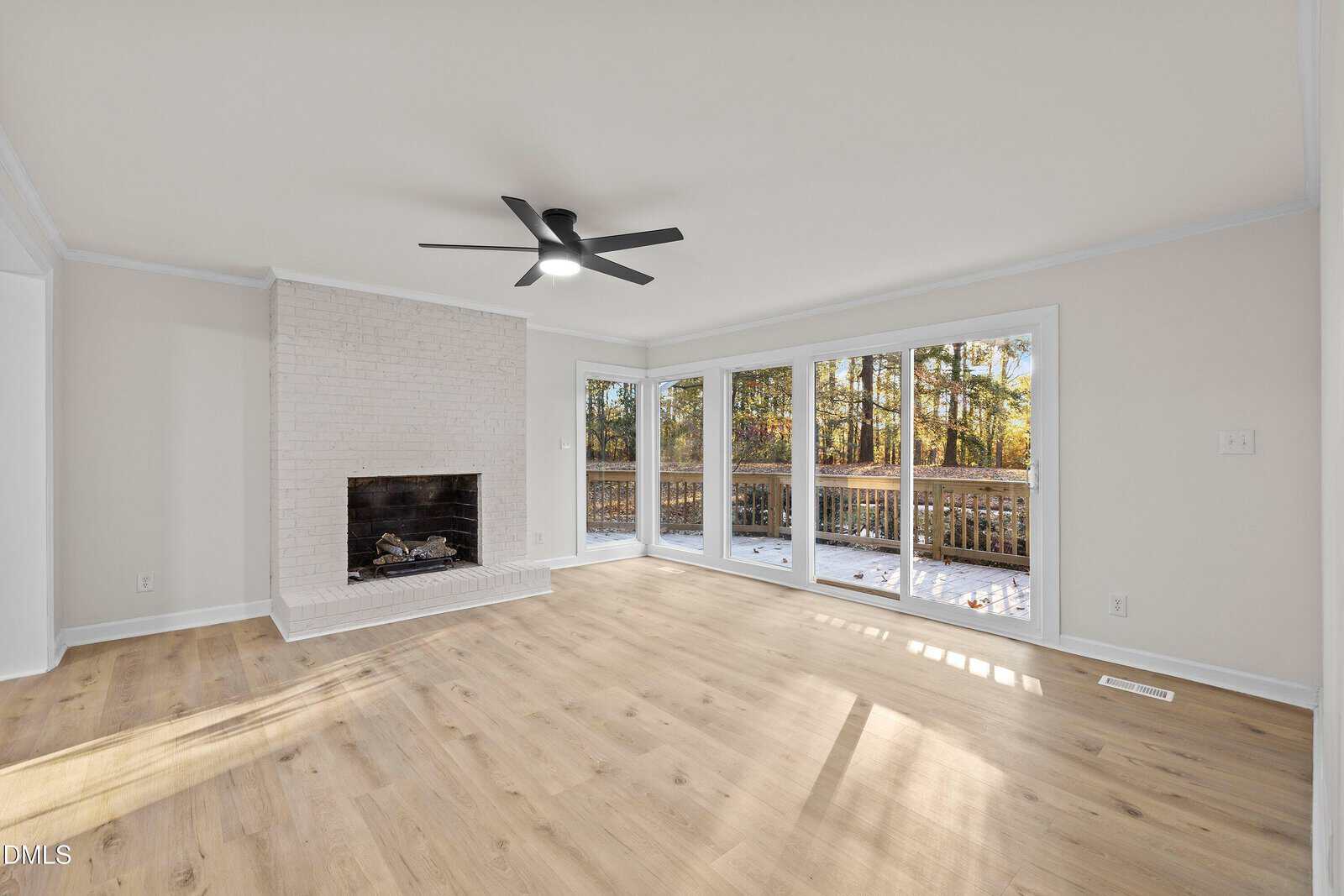 Bright living room with light wood floors, a white brick fireplace, black ceiling fan, and large windows overlooking a deck and wooded area. Sunlight streams in, creating a warm and inviting atmosphere.