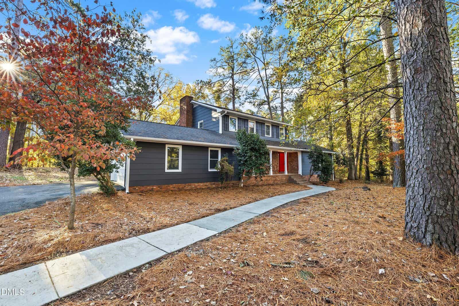 Two-story, gray house with white trim and a red front door, surrounded by tall trees and autumn leaves. A concrete walkway leads from the driveway to the entrance. The yard is covered in pine straw.