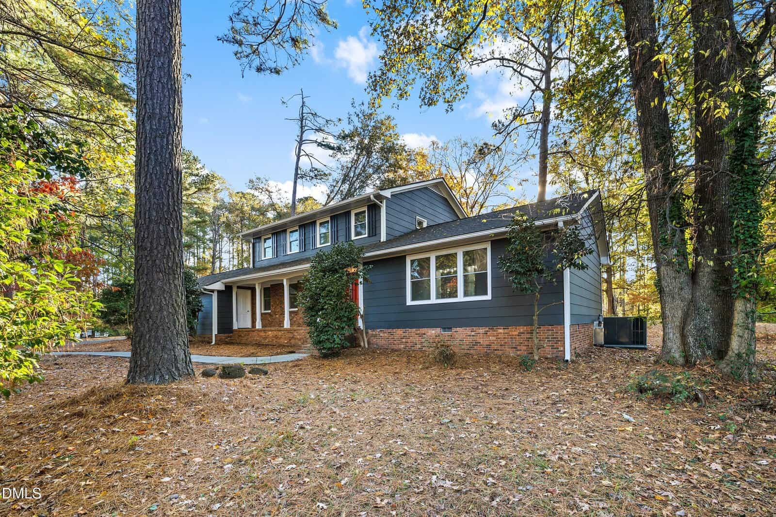 A two-story house with dark gray siding, white trim, and a brick foundation is surrounded by tall trees and fallen pine needles. The home has multiple windows and a covered front porch with columns.