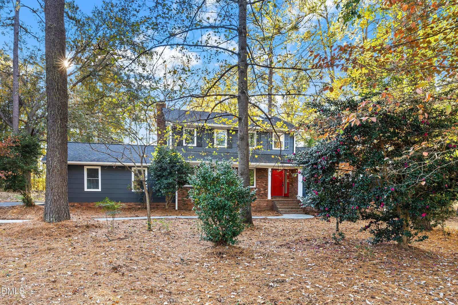 A two-story gray house with white trim and a red front door is surrounded by tall trees and a yard covered with dry pine needles and fallen leaves on a sunny day.