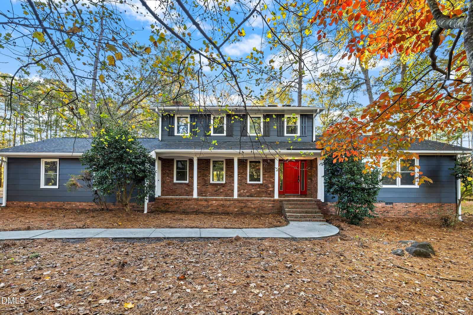 Charming two-story house at 109 Dogwood Circle, Butner, with dark blue-gray siding, red double front doors, brick steps and base. Surrounded by trees with autumn leaves and a yard of pine needles. Curved walkway leads to the entrance.