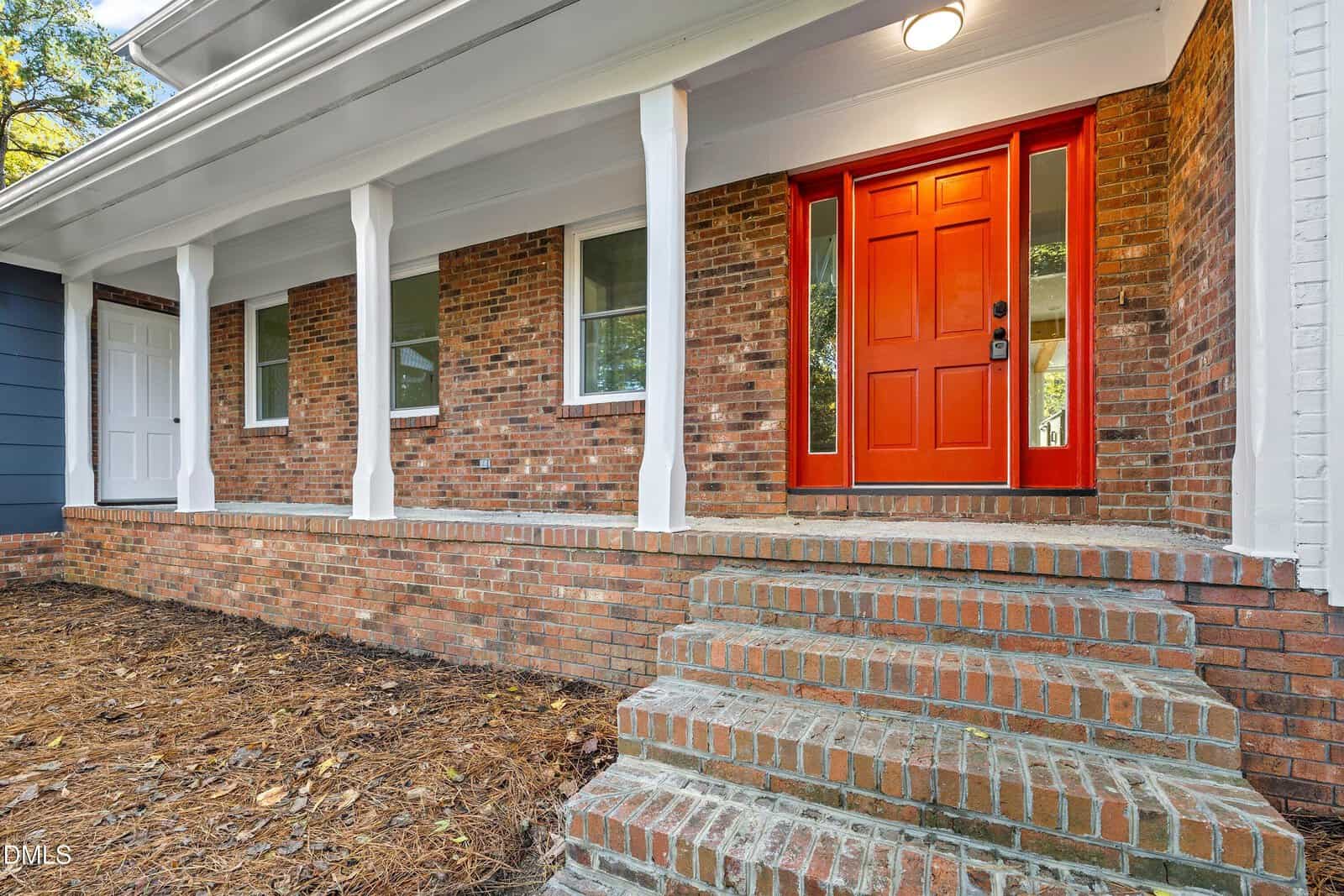 A brick house with a covered front porch, white columns, several windows, and a red front door with glass panels. Brick steps lead up to the entrance. The house is surrounded by mulch and natural landscaping.