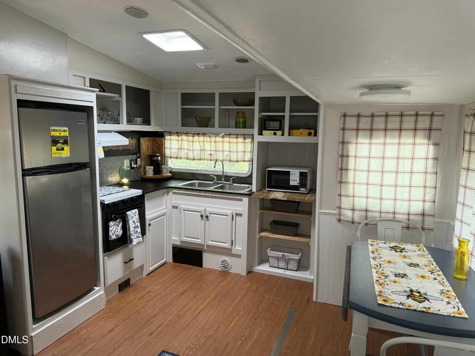 Small kitchen area in a camper or RV features white cabinets, a stainless steel fridge, a stove with a hood, a microwave, and a double sink. A table with bee-themed decor sits near a window with checkered curtains.
