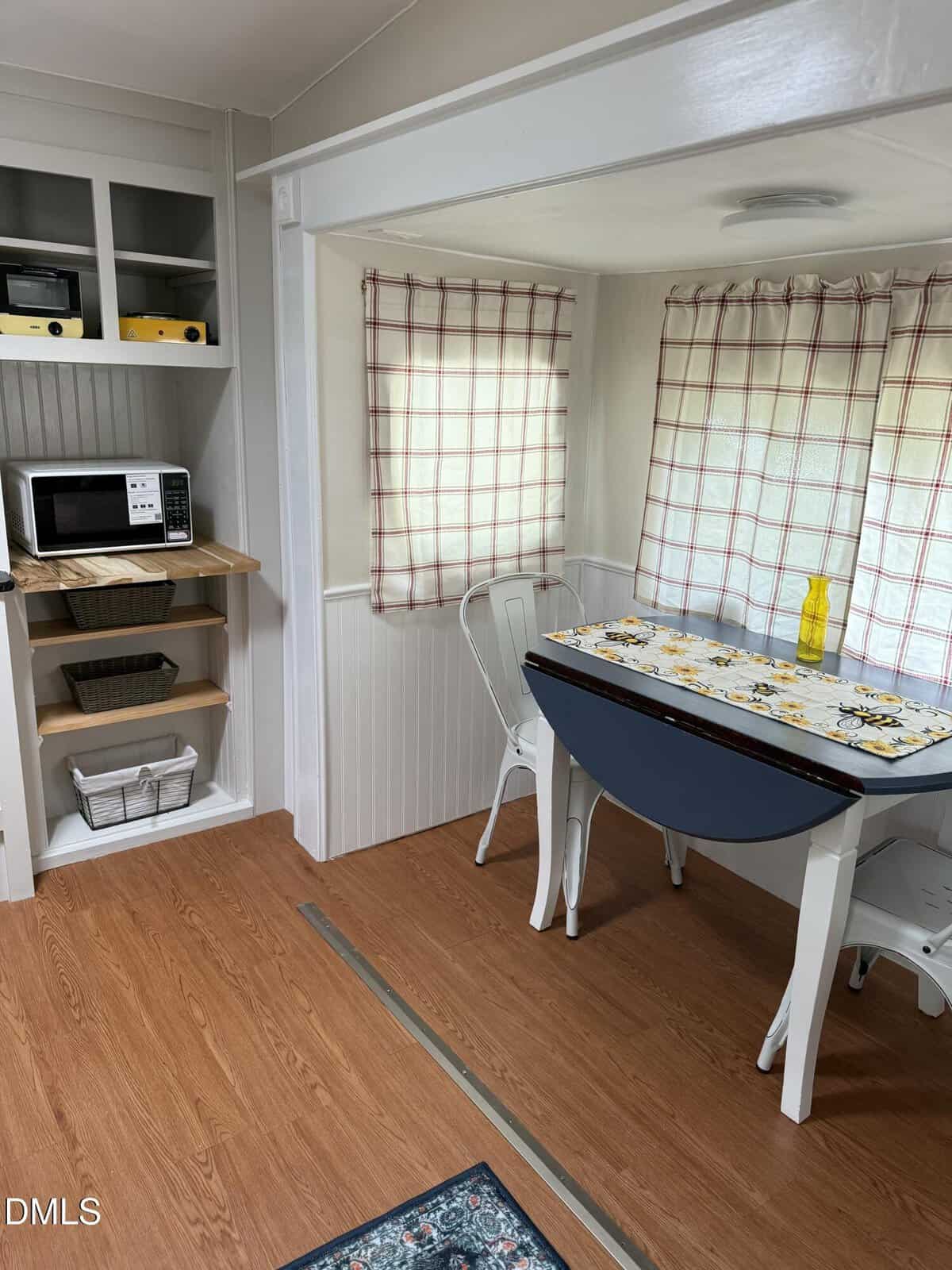 Small dining nook with a drop-leaf table, two white metal chairs, a floral tablecloth, and a yellow vase. Nearby, open shelves hold a microwave, baskets, and small kitchen appliances. Windows have plaid curtains.