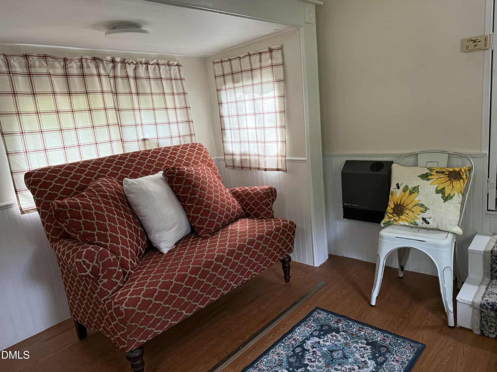 A small room with a red patterned loveseat and two white pillows, a metal chair with a sunflower pillow, plaid curtains on two windows, a small wall heater, hardwood floor, and part of a patterned rug.