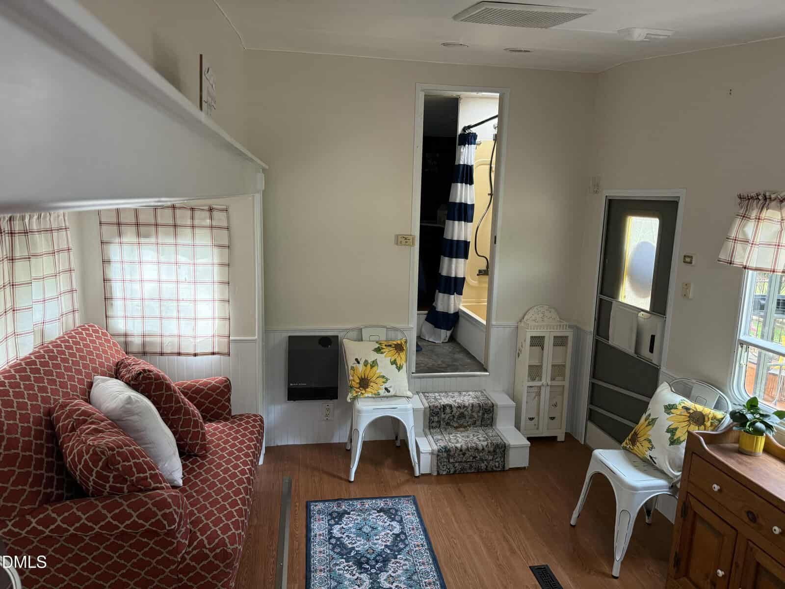 A small living space with a red patterned couch, white chairs with sunflower pillows, a blue rug, and a set of stairs leading to another room with a blue and white striped shower curtain. Light streams in from curtained windows.