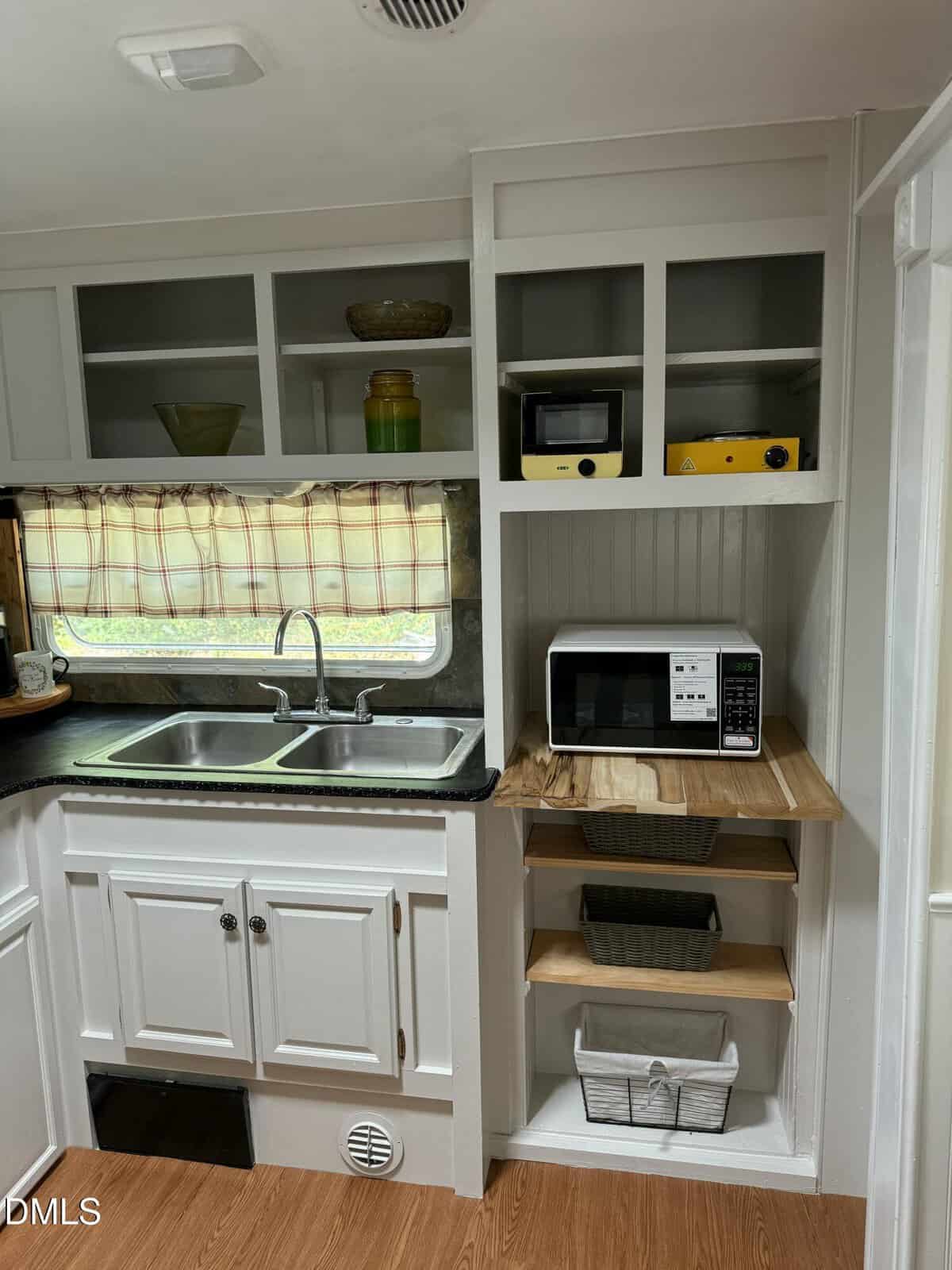 A small kitchen with white cabinets, open shelves holding various containers, a double sink, a microwave on a wooden shelf, and a yellow appliance on an upper shelf. The window has a plaid curtain.