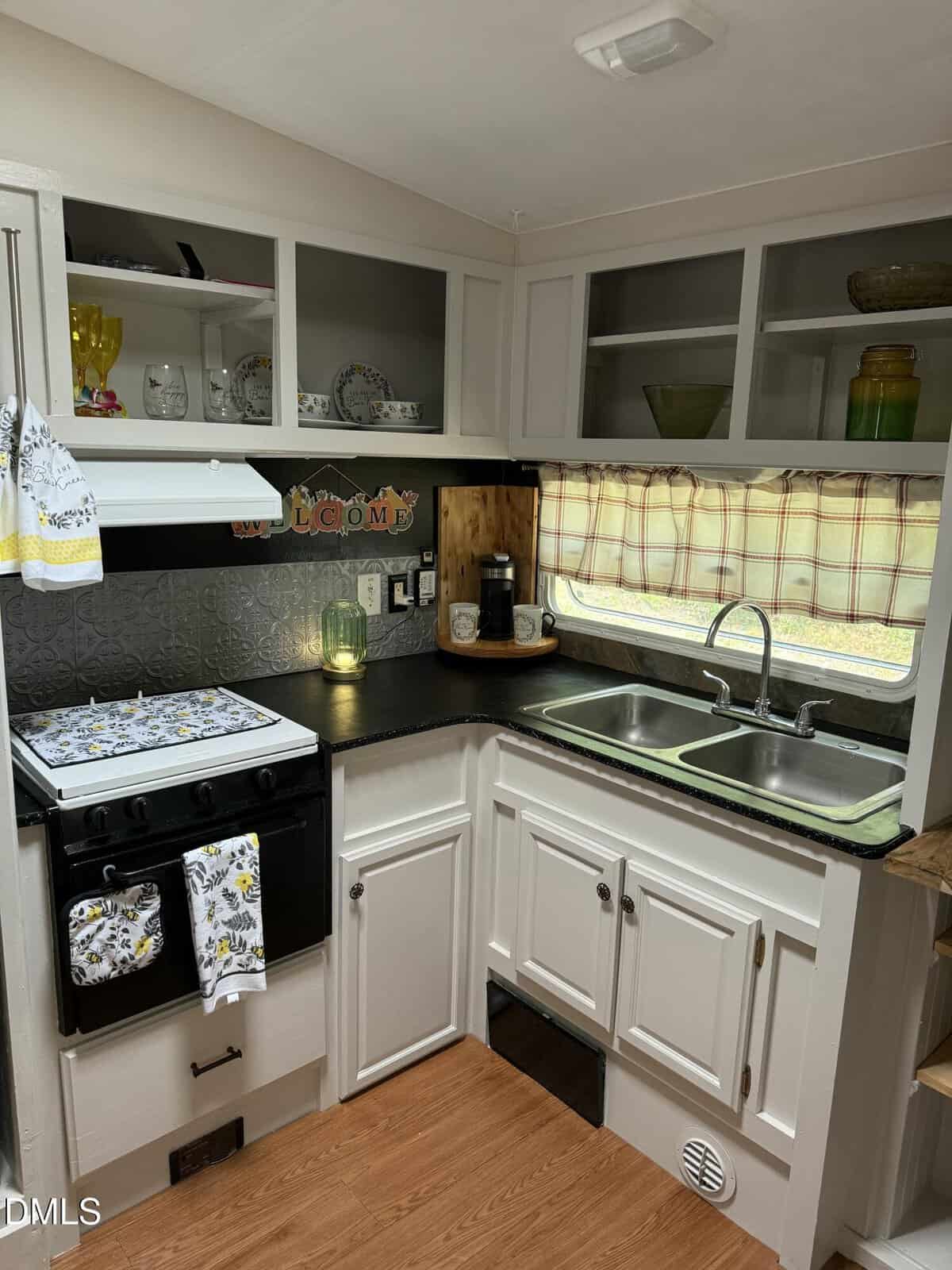 A small kitchen features white cabinets, a stove with a floral towel, open shelves with dishes, a double sink under a window with a plaid curtain, and various decorative items on the counter and shelves.