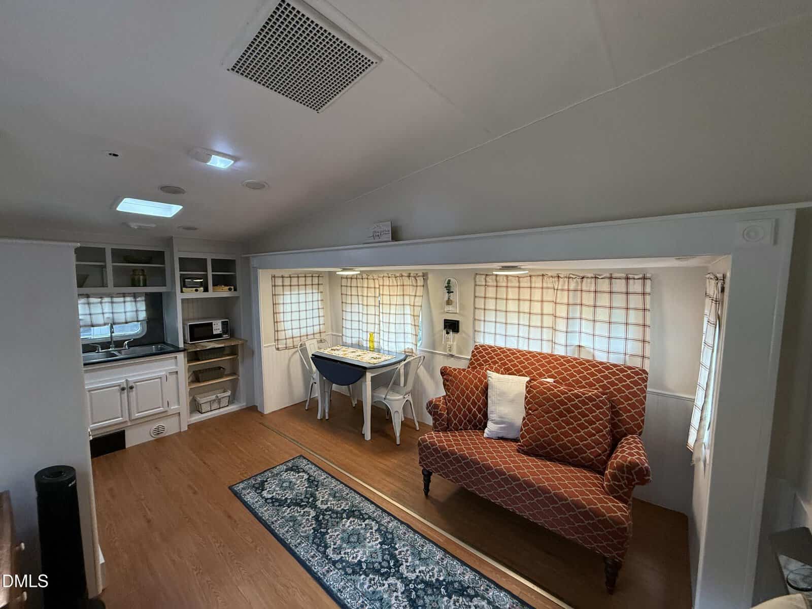 Small living area inside a mobile home or RV, featuring a red patterned loveseat, a dining table with two chairs, a blue rug, wood flooring, and a compact kitchen area with cabinets, a microwave, and a small sink.