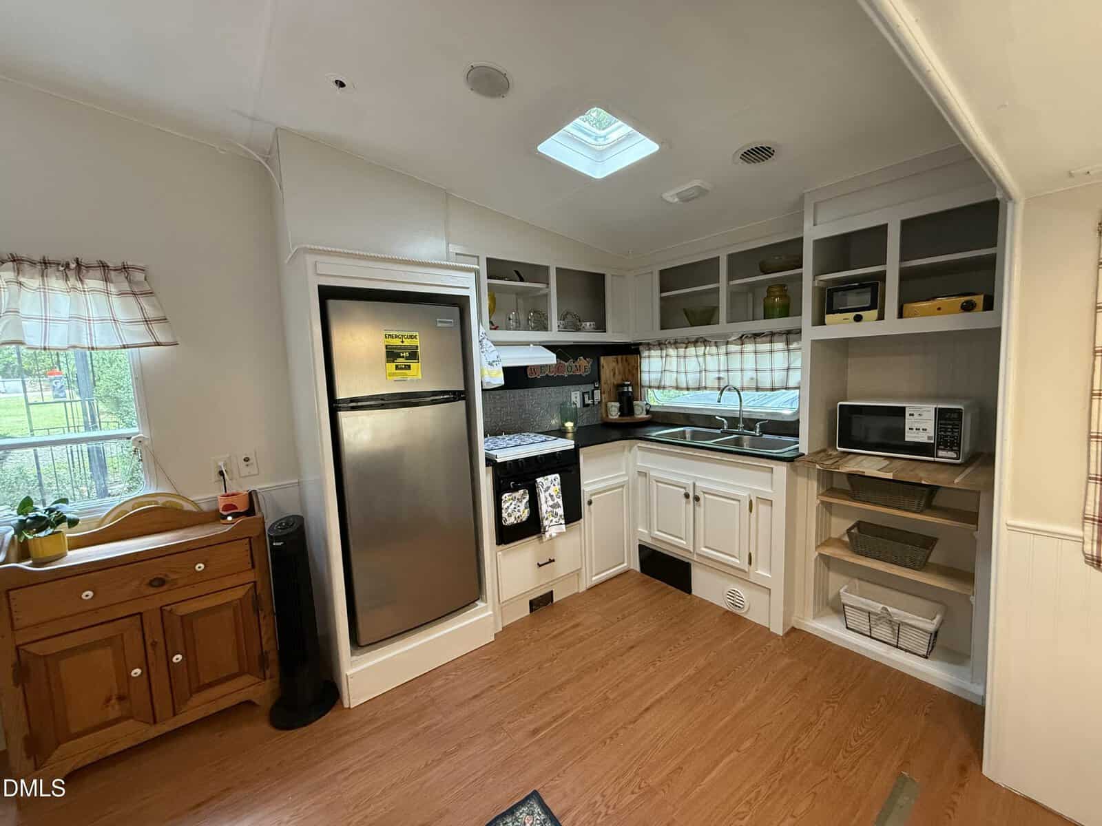 A small kitchen with white cabinets, a stainless steel fridge, stove, microwave, toaster oven, and double sink under a window with checkered curtains. Wooden floors and shelves with various kitchen items are visible.
