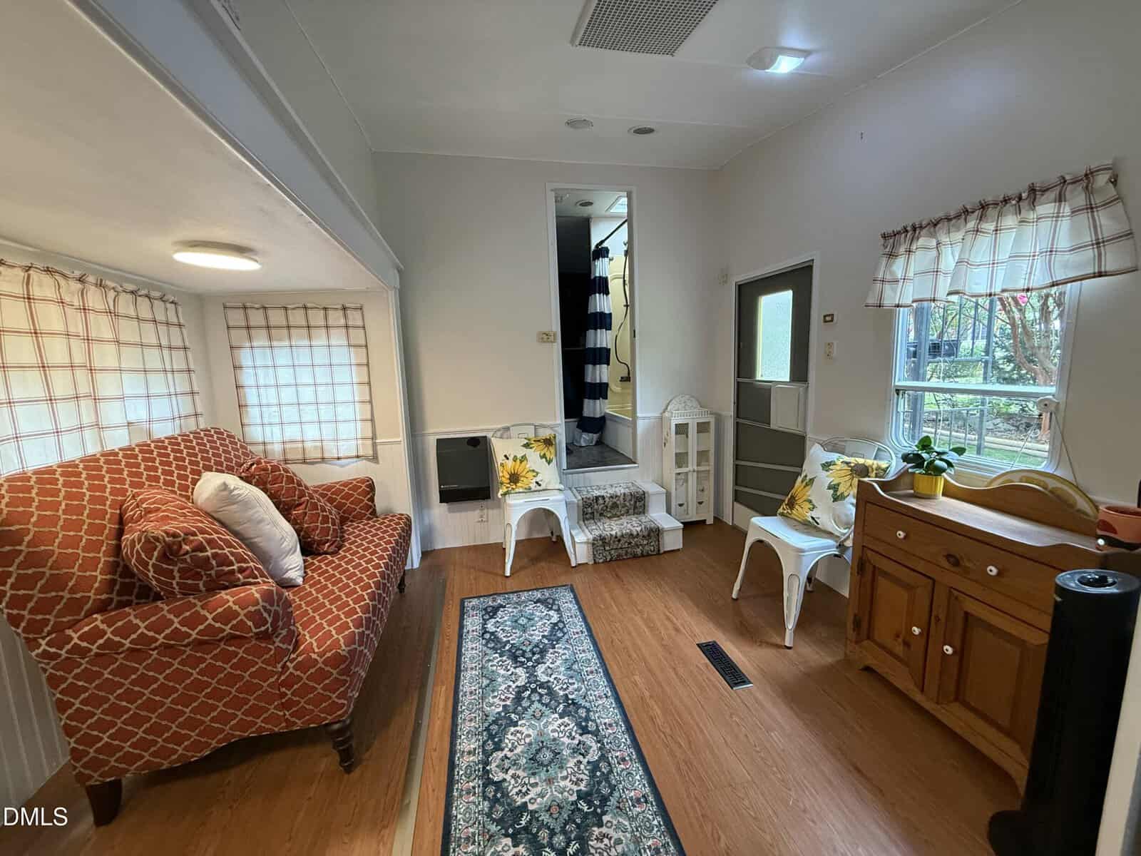 Cozy living area with an orange-patterned armchair, small tables, a wooden sideboard, potted plants, checkered curtains, and a narrow runner rug. Steps lead to an adjoining room with striped shower curtain visible.