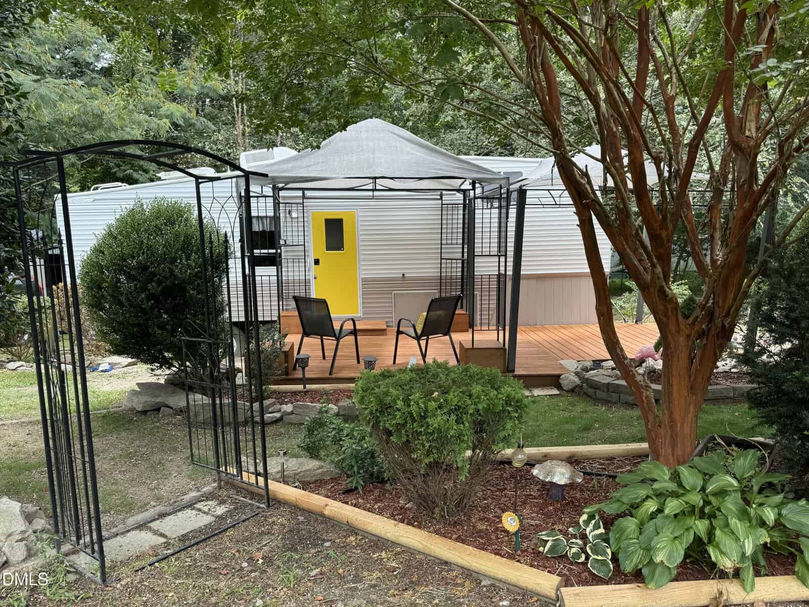 A small trailer with a bright yellow door sits on a wooden deck, surrounded by trees and bushes. Two chairs and a small table are arranged on the deck, and a metal archway leads to the entrance.