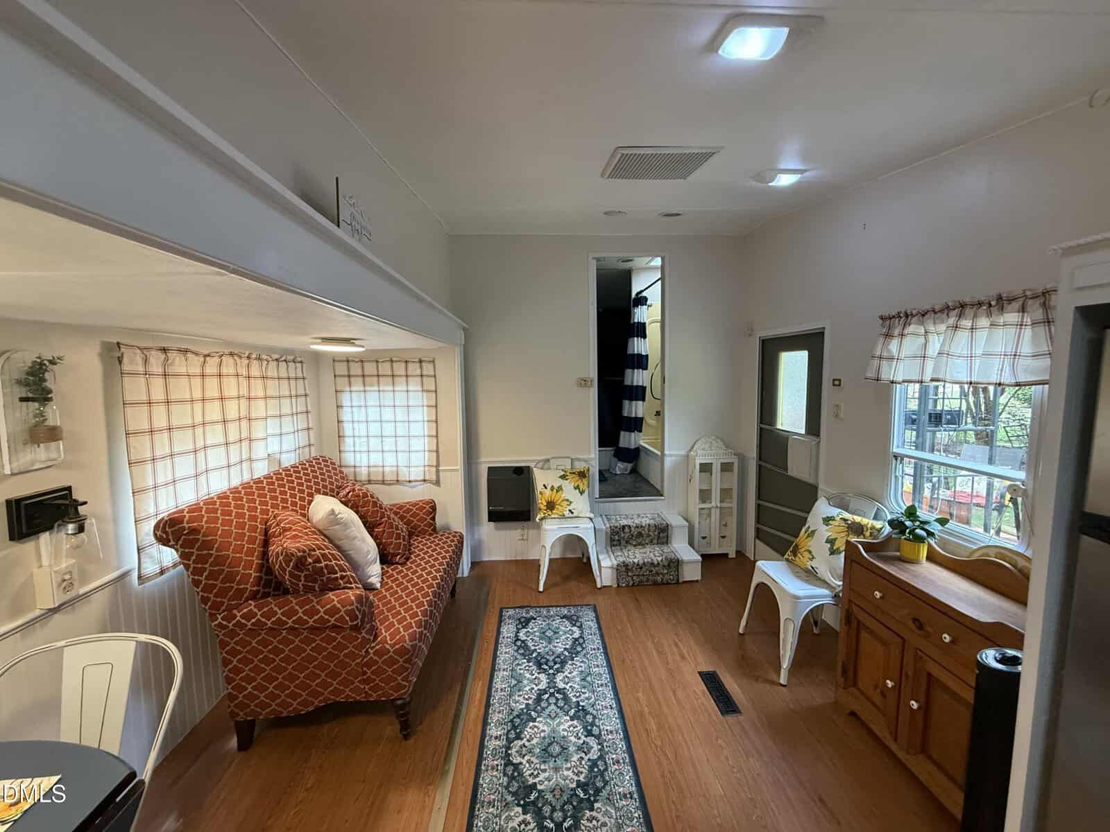 Cozy living room with a red patterned chair, floral pillows, wooden floor, rug, plaid curtains, white furniture, small table, potted plants, and steps leading to a raised area with a striped shower curtain visible.
