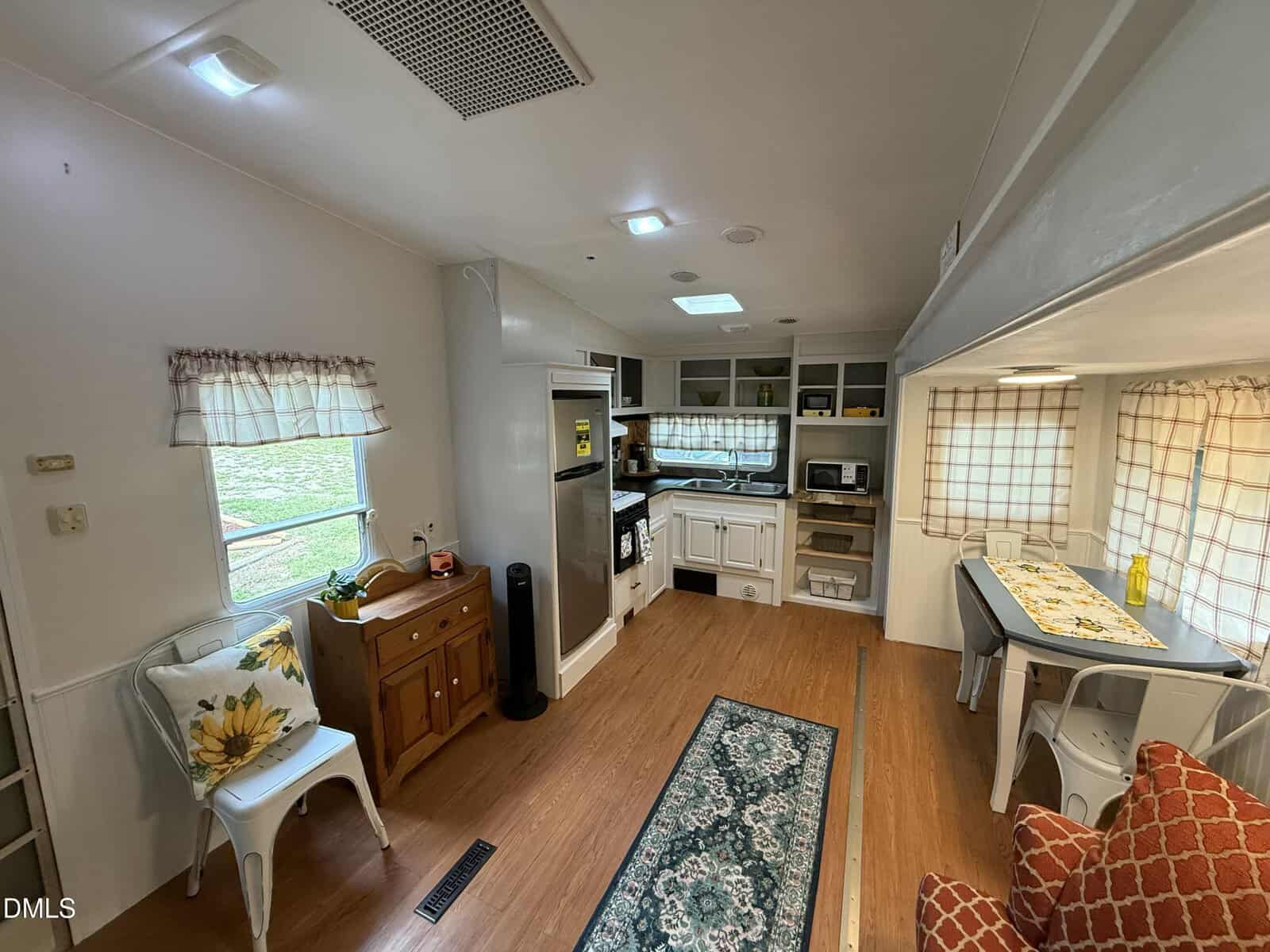 A cozy RV interior with wood floors, a kitchen featuring white cabinets and appliances, a dining area with a sunflower pillow, and large windows with plaid curtains letting in natural light.