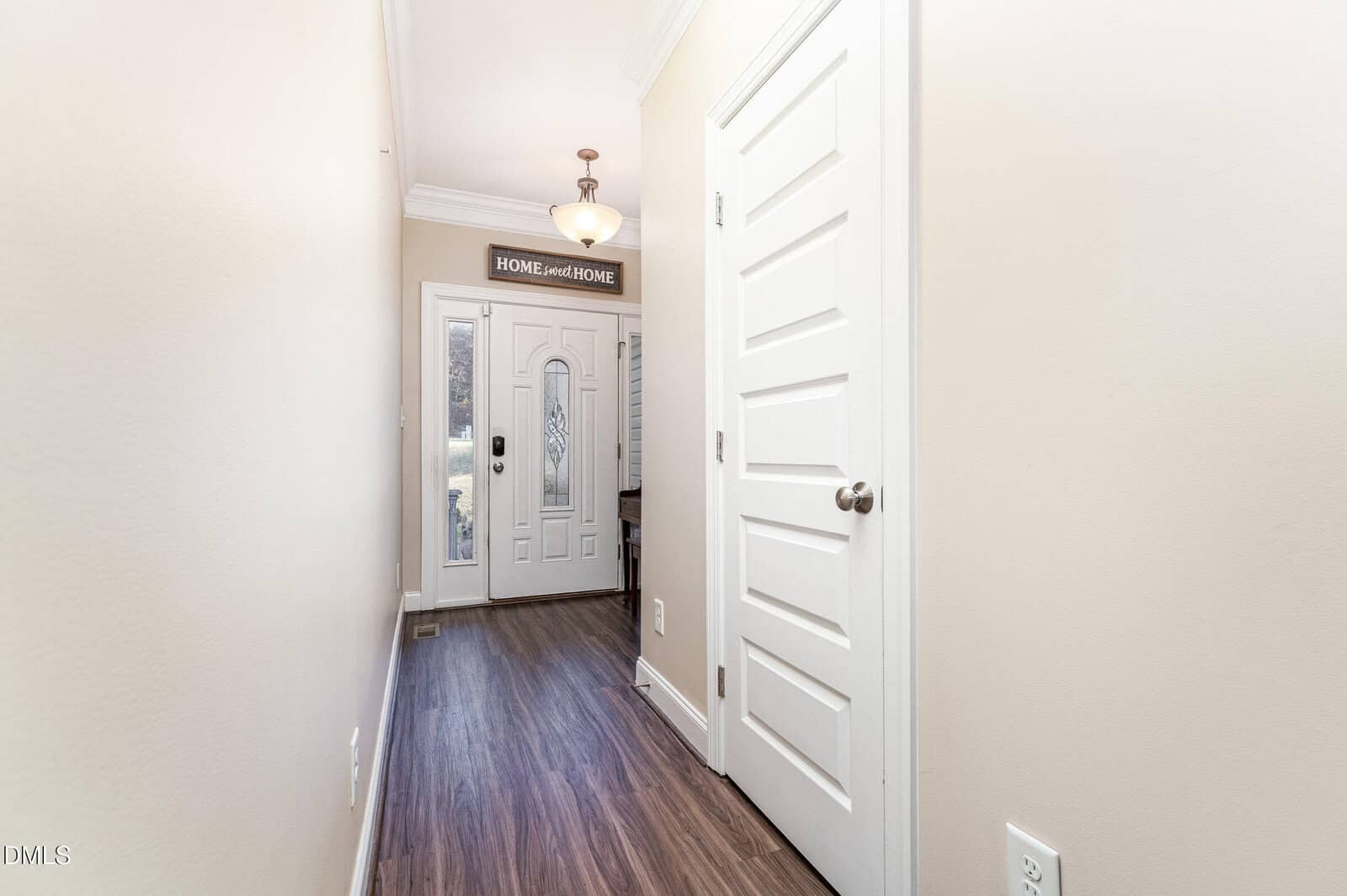 A hallway with light-colored walls and wood flooring leads to a front door with glass panels. A ceiling light hangs above, and a sign above the door reads "HOME sweet HOME." Several doors are on the right side of the hall.