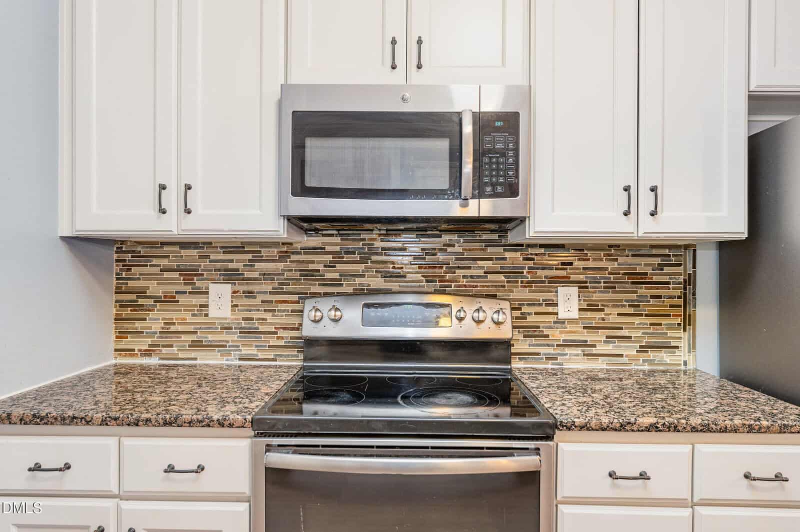 A modern kitchen features a stainless steel oven with a matching microwave above it, white cabinets, granite countertops, and a multicolored tile backsplash.