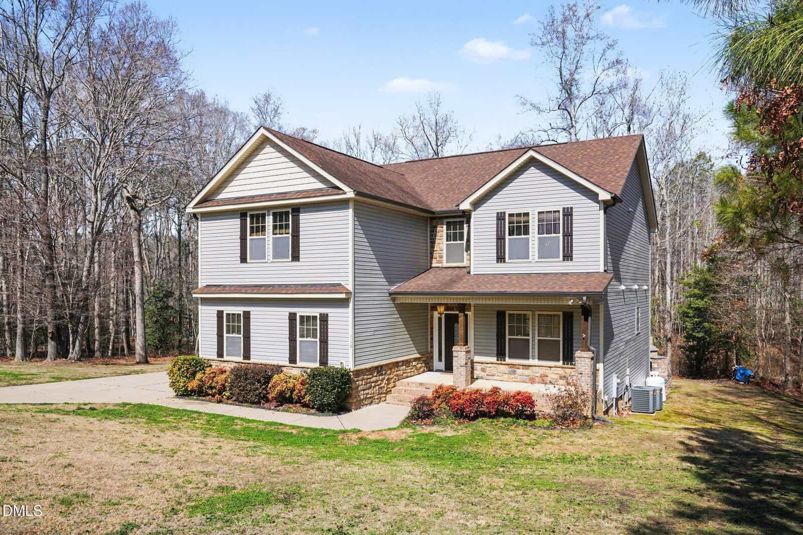 Two-story house with gray siding, brown roof, and stone accents on lower facade. The house has multiple windows and a covered front porch. It is surrounded by a grassy lawn and trees in the background.