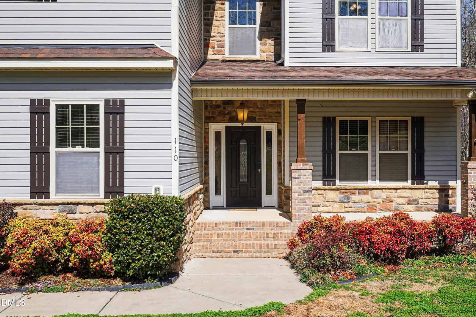 Front view of a house with light-colored siding, stone accents, dark shutters, a covered porch, and a black front door. Shrubs and colorful bushes line the walkway leading to the entrance.
