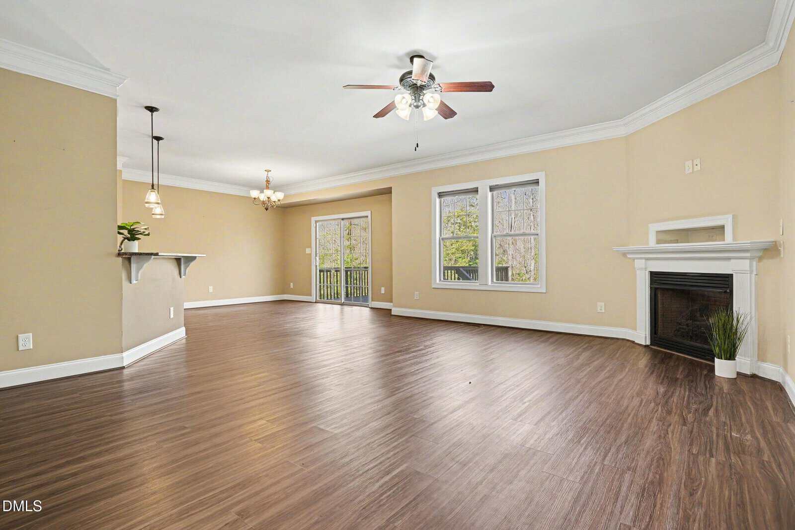 Spacious empty living room with beige walls, white trim, dark wood floors, ceiling fan, fireplace, large windows, sliding glass door, and open connection to dining area with two light fixtures.