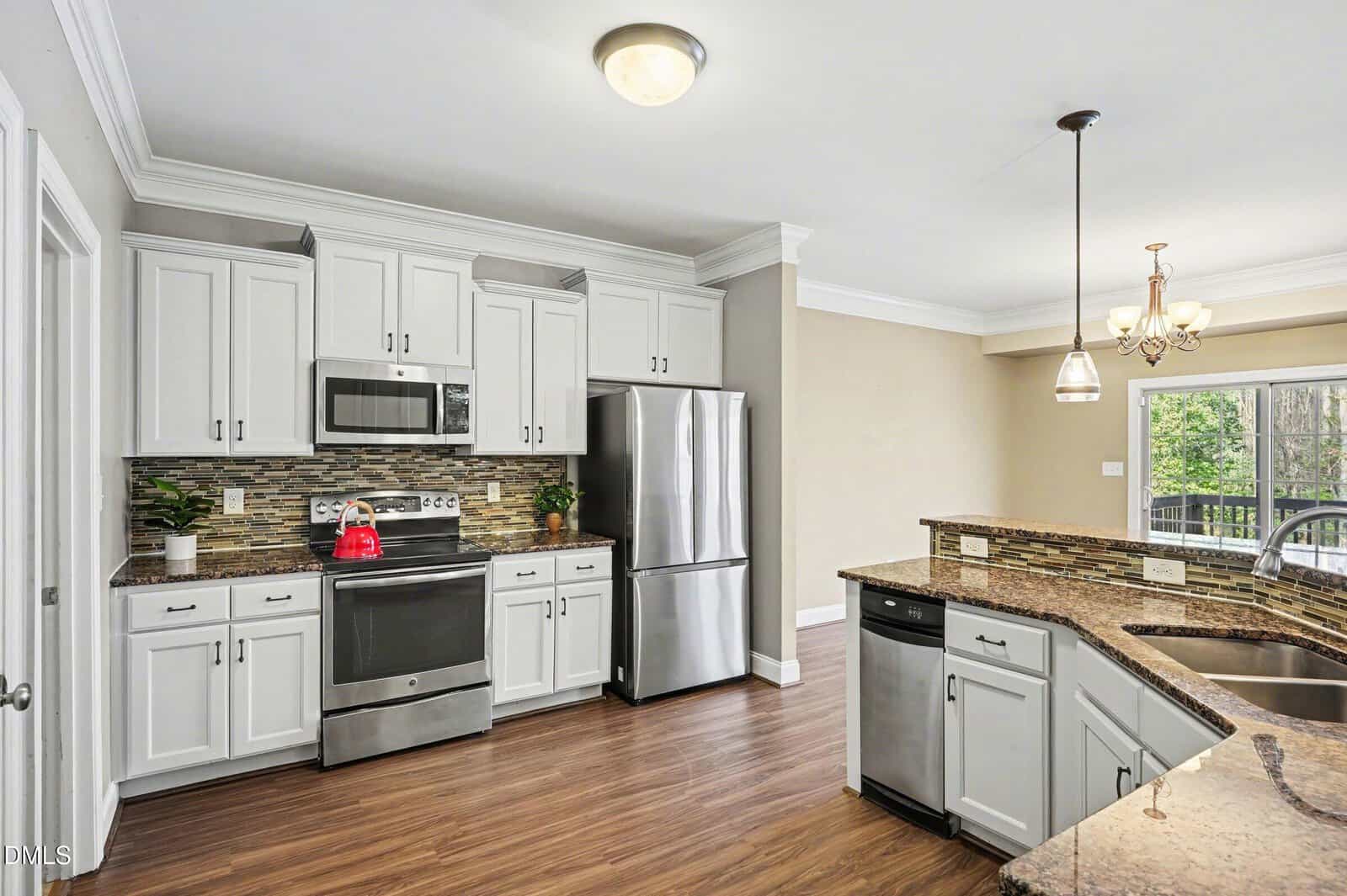 Modern kitchen with white cabinets, stainless steel appliances, a tile backsplash, and wood flooring. There is a sink and dishwasher in the granite island, with pendant lighting and a window in the adjacent dining area.